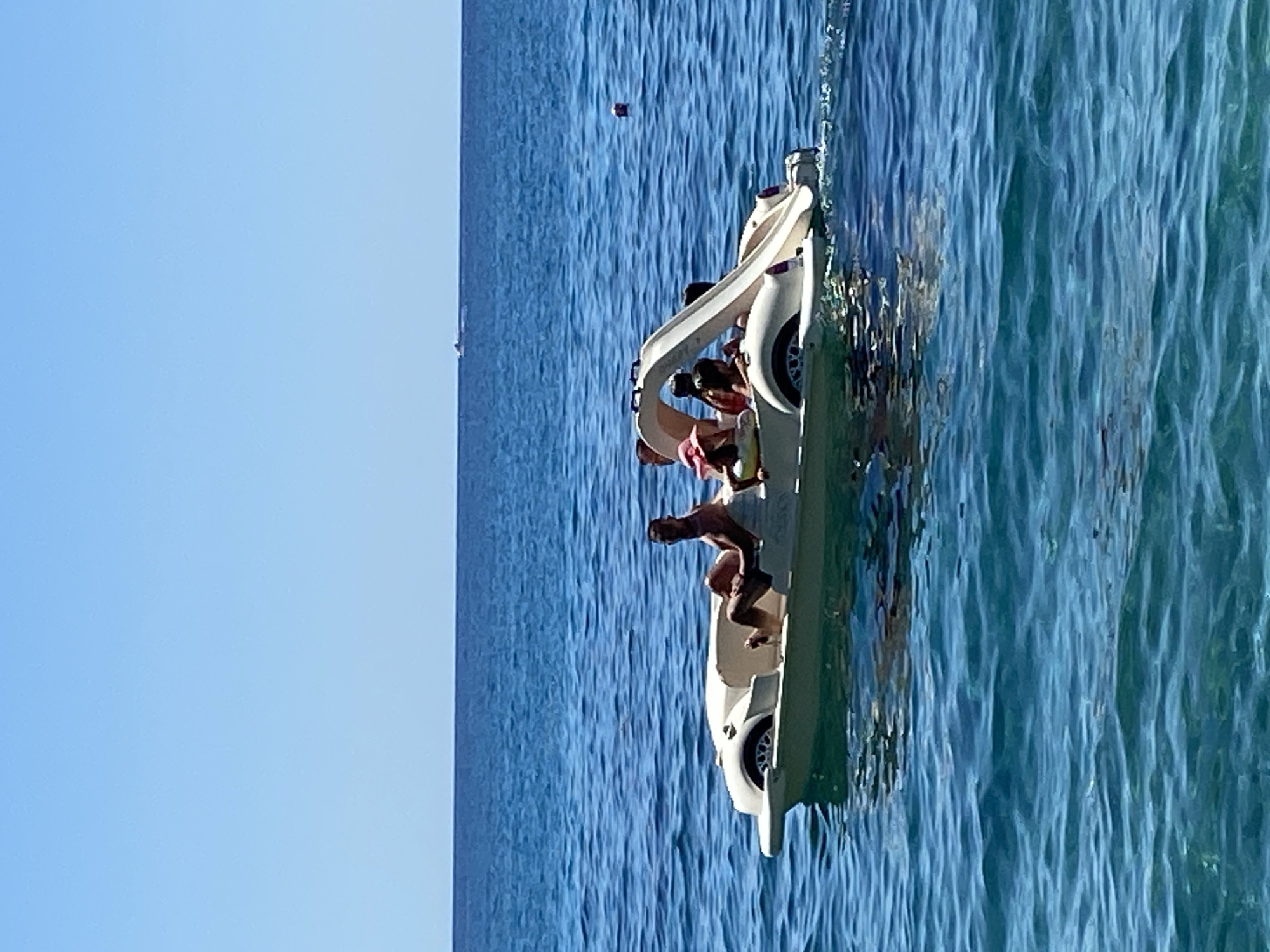 A pedal boat shaped like a vintage car is on the water. The boat is light-colored and has four people on board. One person is in the front, appearing to be pedaling. Two people are in the middle, and one person is in the back. The water is a clear blue, and the sky is a bright, cloudless blue. In the distance, there is a faint outline of land and another small boat. The scene suggests a sunny day with people enjoying a recreational activity on the water. The location is not specified in the image.