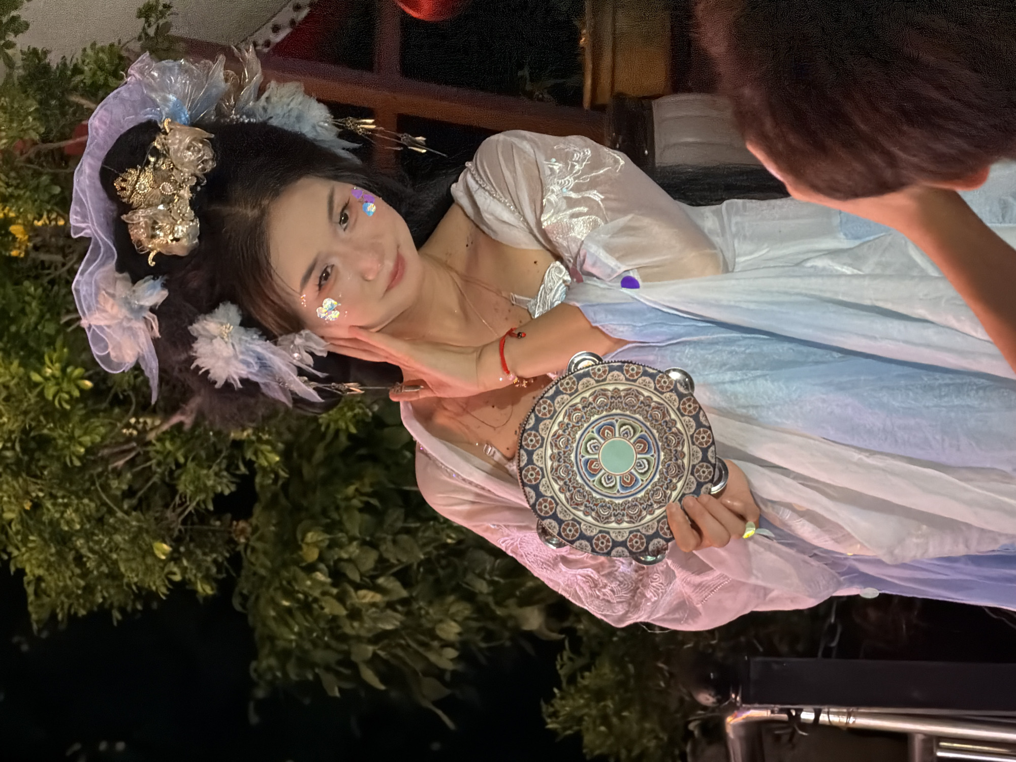 A young East Asian woman with dark hair styled in an elaborate updo adorned with blue and white ornaments and a golden crown is shown in a close-up portrait. She wears a white and light blue flowing costume with sheer sleeves and shimmering embellishments. Her face is decorated with iridescent glitter. She holds a round, decorative tambourine with a mandala-like pattern in her right hand, touching her neck with her left. Her gaze is directed slightly away from the camera with a subtle smile. The background features lush green foliage and a dark, indistinct environment, suggesting an outdoor setting at night. A portion of another person's head and shoulder is visible in the lower right corner. The scene appears to be a staged performance or celebration.