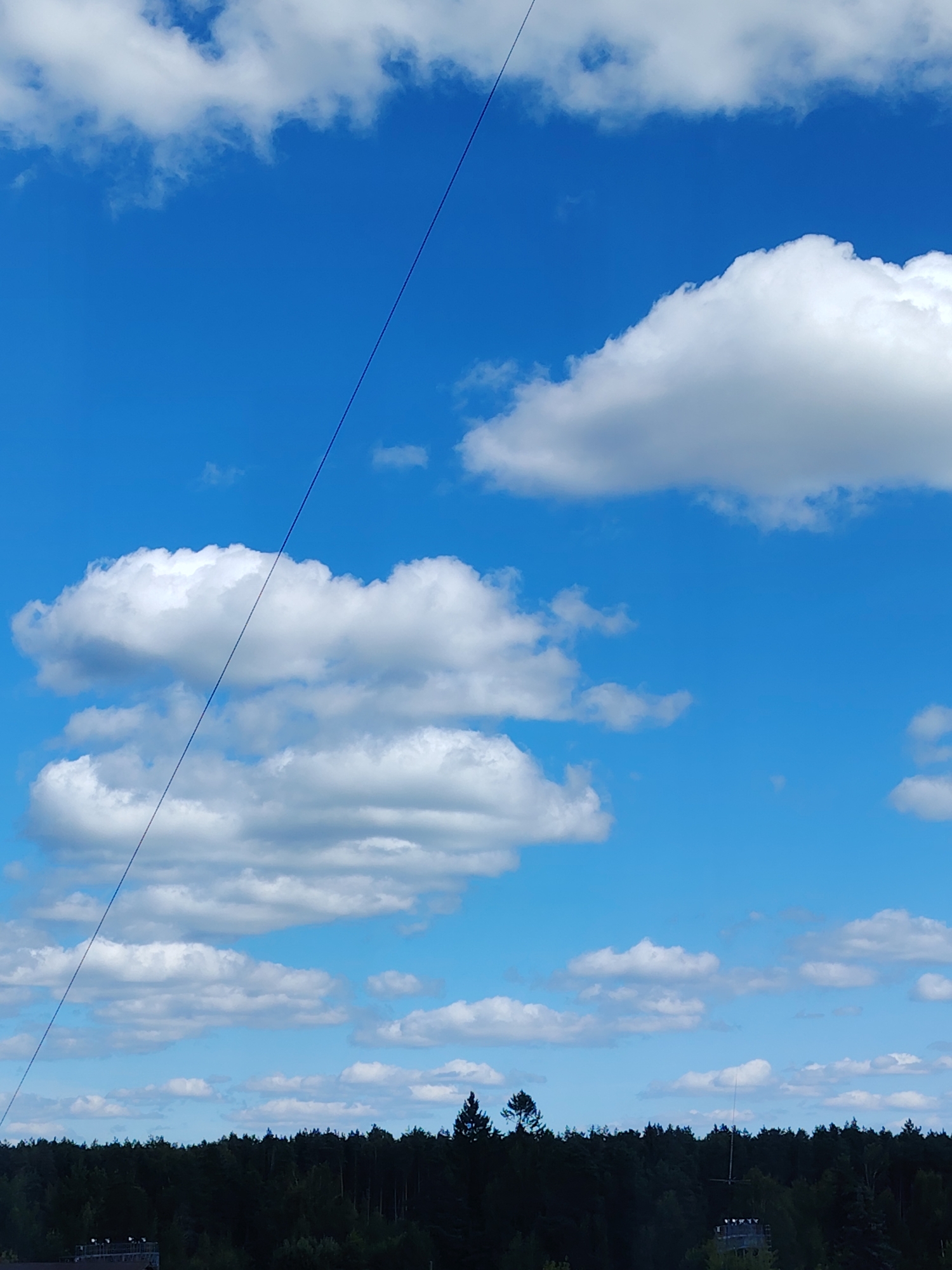 A bright, sunny day is depicted with a vibrant blue sky filled with scattered white cumulus clouds. A thin, dark line, likely a wire or cable, diagonally bisects the sky from the upper left to the middle right. Below the sky is a dense, dark green line of trees forming a forest canopy that stretches across the horizon. Visible above the trees on the far right is a single, dark green coniferous tree with a pointed top. In the lower left corner, partially obscured by the treeline, are the tops of some man-made structures, possibly buildings or towers, with visible horizontal railings and lights. The overall scene suggests an outdoor environment, likely in a rural or semi-rural area. The presence of the wire indicates some form of infrastructure, such as power lines or a zipline, is present in the vicinity of Konakovo, Russia.