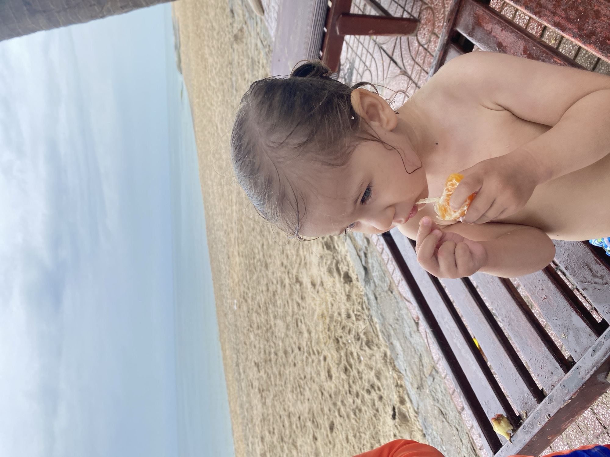 A child eats an orange while sitting on a wet, wooden bench. In the background is a beach landscape of Phu Thuy Ward, Vietnam, featuring a sand-covered area, the ocean, and an overcast sky.