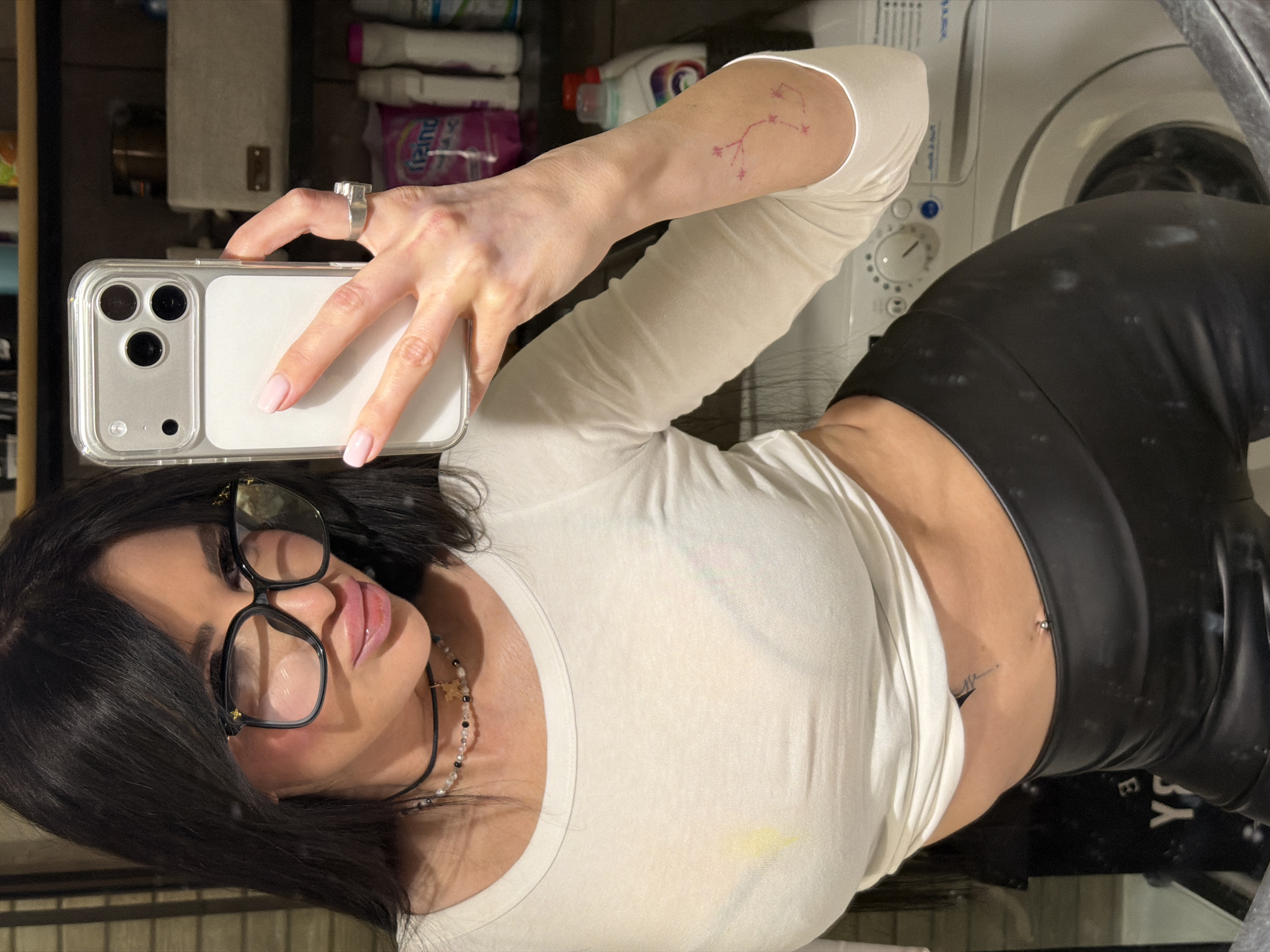 The image is a mirror selfie taken by a woman indoors, rotated 90 degrees counter-clockwise. The setting appears to be a utility or laundry room, likely in a residence in Moscow, Russia.
**Subjects:**
The central subject is a woman with dark hair, parted and styled, wearing large black-framed prescription glasses. She has makeup, including glossy pink lips. She is wearing a light-colored (possibly off-white) long-sleeved crop top and dark, likely leather or pleather, high-waisted pants. Visible accessories include a silver-toned, chunky ring on her right index finger and a necklace made of black beads, white pearls, and a small gold-colored charm. She has a belly button piercing and two tattoos: a small, red constellation-like design on her right inner bicep/forearm, and another small, dark, abstract tattoo resembling a heartbeat on her lower left abdomen. Her fingernails are manicured with light pink polish. She is holding a silver/white iPhone in a clear protective case, featuring a triple-lens camera array, in her right hand.
**Objects:**
In the background, a white front-loading washing machine is visible to the right. Above it, various cleaning products and bottles, some in pink packaging (e.g., "Lenor Unstoppables"), are stored on shelves.
**Activity or event taking place:**
The woman is taking a mirror selfie.
**Notable details:**
The woman has a neutral to slightly alluring expression. There is a faint yellow stain on the front of her crop top. The scene is illuminated by indoor lighting, and no specific time of day or weather conditions can be discerned. The context provided specifies the location as Moscow, Russia.
**Visible text:**
Text on product packaging in the background, such as "Lenor Unstoppables," is partially visible. Some unreadable text is also present on the washing machine's control panel.