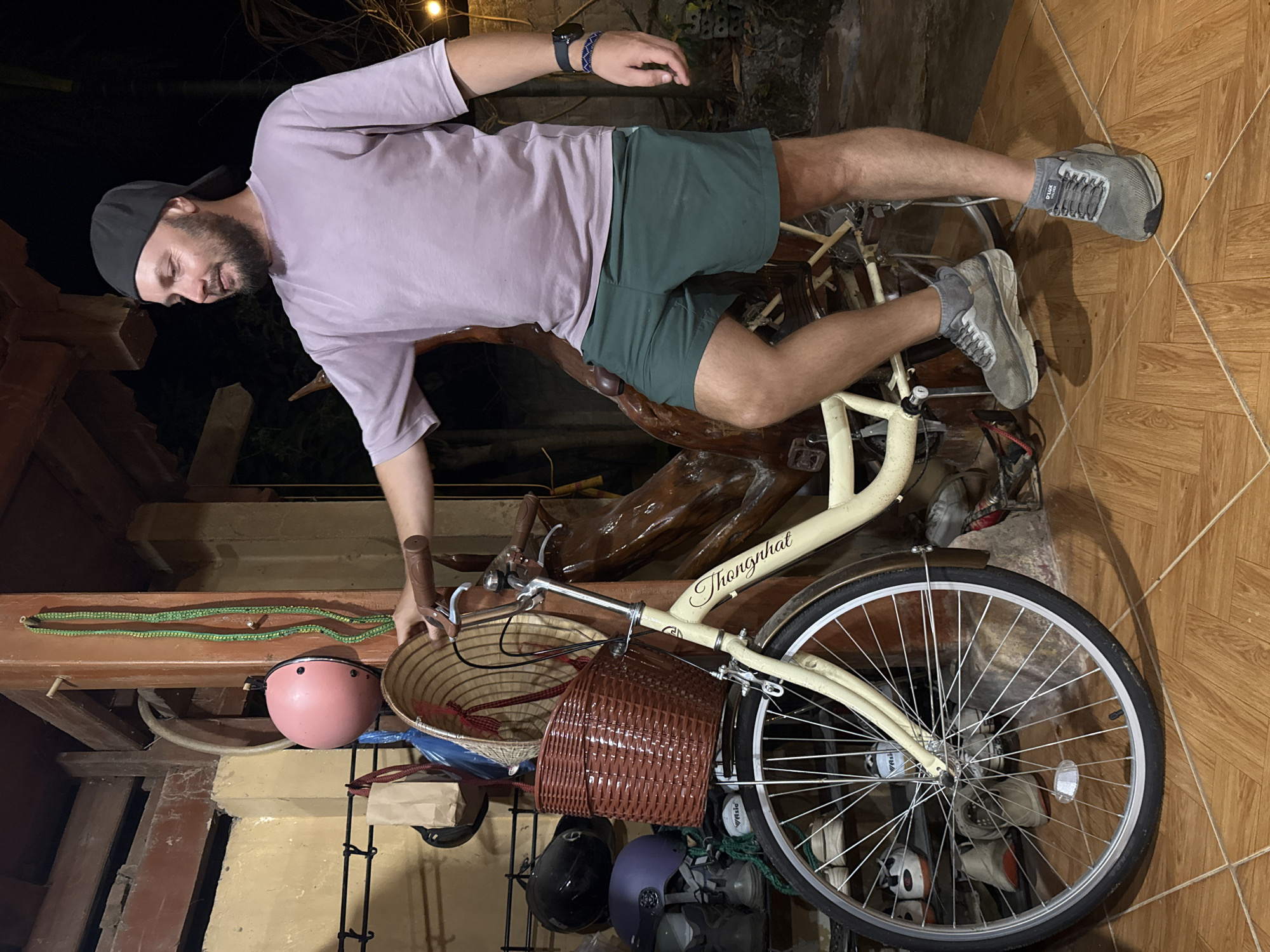 A person is standing next to a bicycle indoors. The bicycle is off-white with a basket on the front. There is a pink helmet and other items hanging nearby. The person is wearing a light purple shirt, green shorts, grey socks, and grey shoes. The location is in the city of Hà Giang 1 Ward, Vietnam.