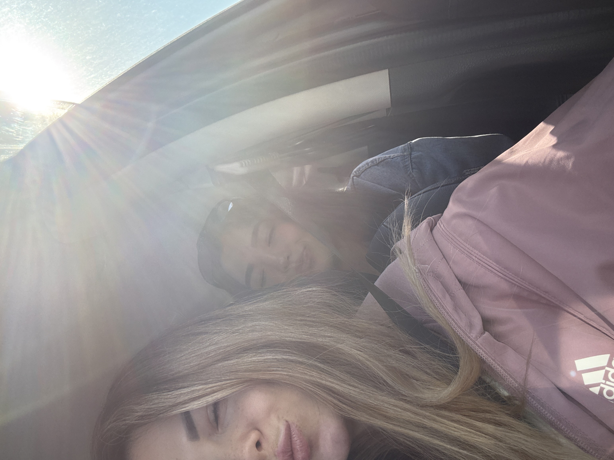Two young women are seen resting or sleeping inside a vehicle, illuminated by bright sunlight streaming through a window. The image is taken from a low angle, looking upwards towards the vehicle's ceiling and window.
In the foreground, a woman with long, light blonde hair is visible, her eyes closed and lips slightly parted. She is wearing a pink or mauve athletic top, on which the white "Adidas" logo is clearly visible on the lower right.
Behind her, another woman is partially visible, also with closed eyes. She has darker hair and appears to be wearing a blue denim jacket.
Strong sunlight enters from the top left of the frame, creating prominent lens flare and light rays that brighten the scene and cast a soft, hazy glow. The sun's brightness suggests it is daytime. The interior of the vehicle, including parts of the headliner and window frame, is discernible. Both women appear to be in a state of peaceful rest.