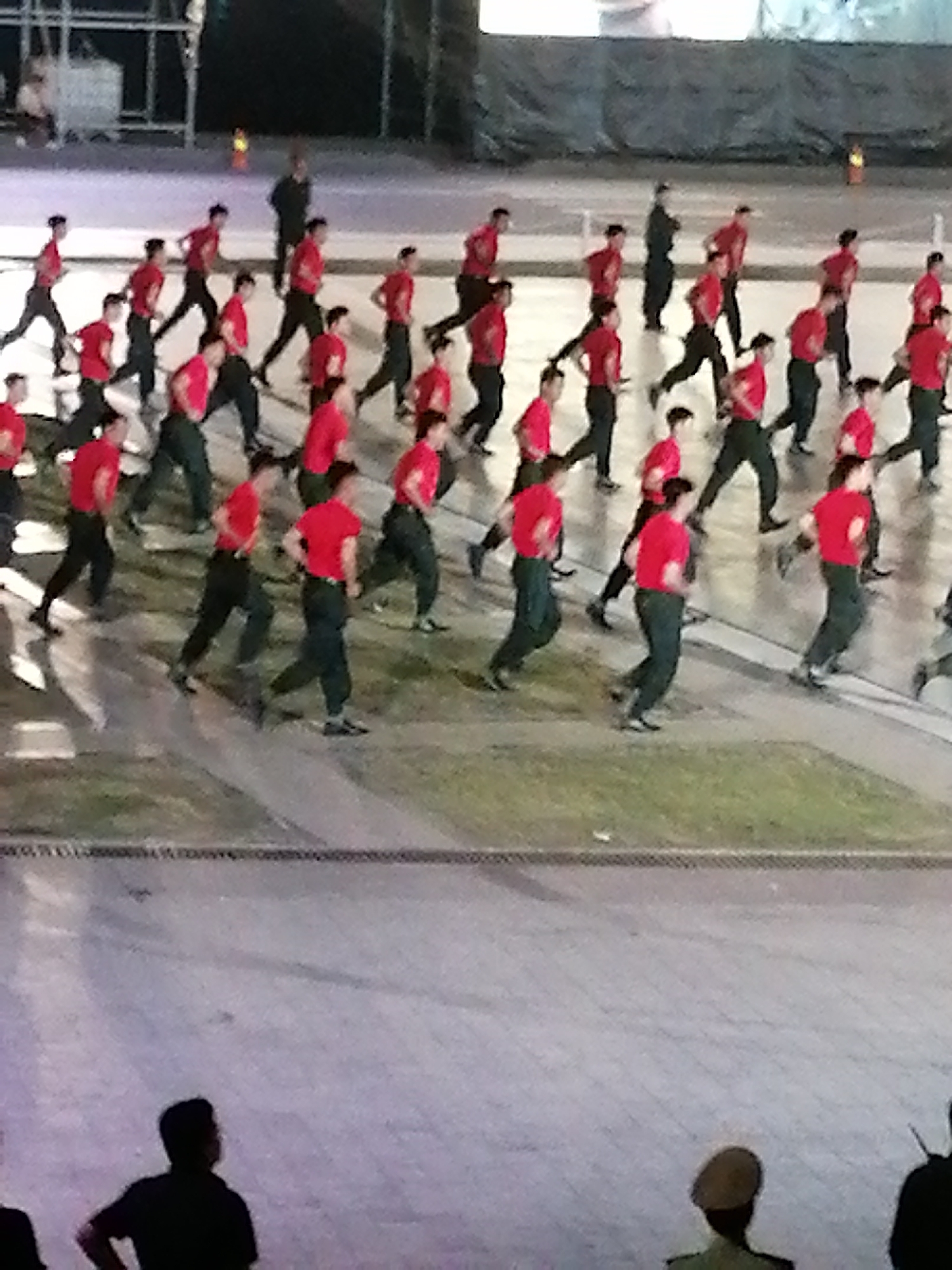 The media file depicts a group of people, predominantly young men, engaged in a synchronized movement, possibly a march or a drill, on a paved surface. They are dressed in matching outfits consisting of bright red t-shirts and dark pants, with some individuals wearing caps. The group is arranged in rows and columns, creating a sense of order and discipline. The activity appears to be taking place outdoors, with a paved area in the foreground and what looks like bleachers or a stage structure in the background, suggesting a performance or a ceremonial event. The lighting suggests it is either late afternoon or early evening. There are a few other individuals present on the periphery, observing the main group. One person in the foreground is wearing a black shirt and is turned away from the camera. Another person is wearing a light-colored cap and a green uniform, also facing away. The scene is somewhat blurred, which could be due to the motion of the subjects or the quality of the image. The overall atmosphere seems formal and organized. No visible text is present in the image. The context provided mentions Da Lat, Vietnam, which could imply the location of this event.