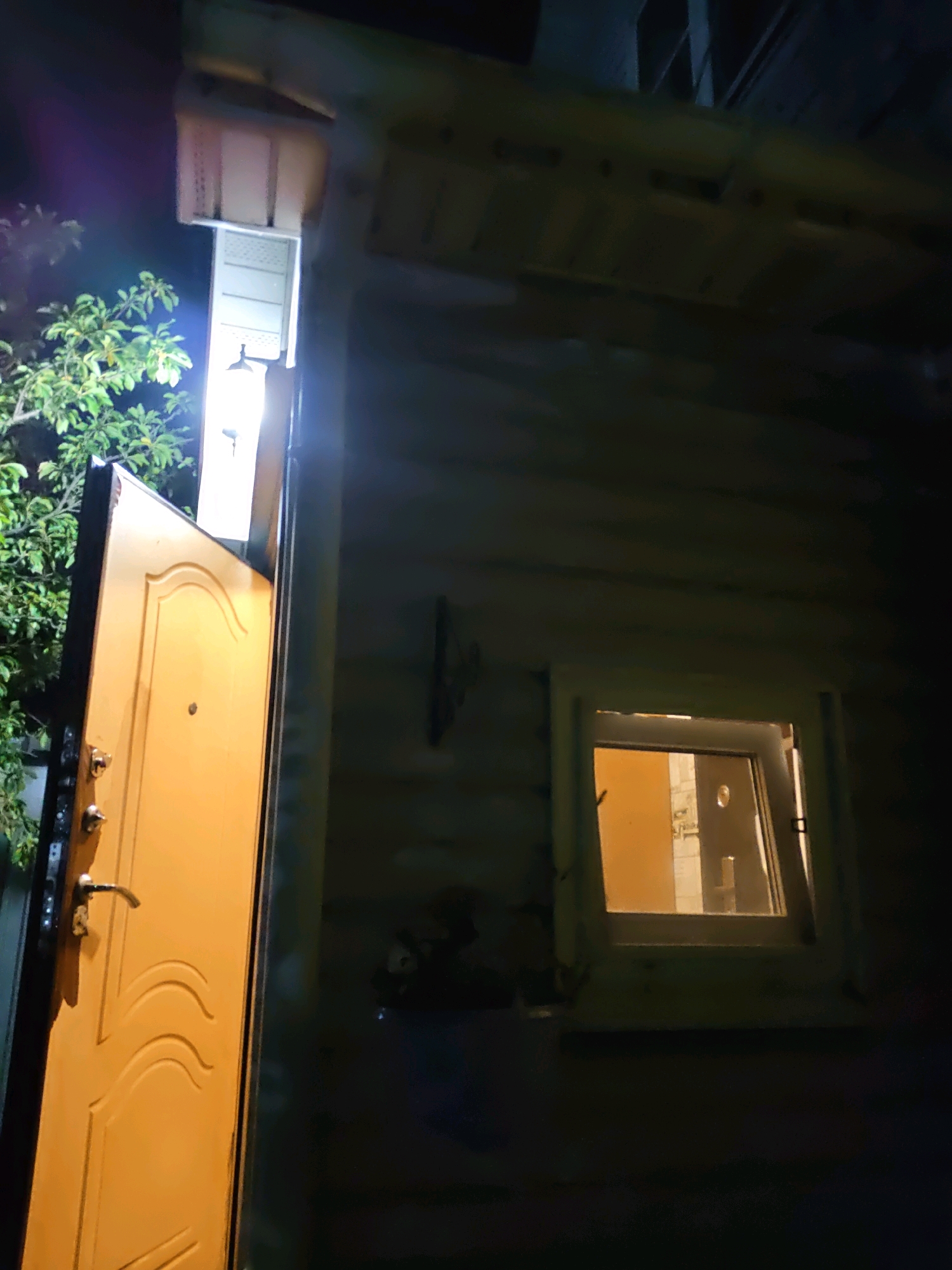 An exterior night shot captures an open, yellow-painted wooden door and a window in a log cabin-style building. The door, positioned on the left, is ajar, revealing a light source within and a key hanging from a hook. To the right, a small, square window glows with warm light, offering a glimpse of interior details, including what appears to be a doorway. The building's log construction is evident, with horizontal wooden planks forming the wall. Overhanging the door is a portion of the roof structure, with exposed wooden beams. To the left of the door, a leafy tree with green foliage is visible, with a bright light source illuminating it from above and casting a purple lens flare into the scene. The ground is not visible. The image is devoid of people. The scene is consistent with the exterior of a dwelling, possibly a bathhouse or guesthouse, in a rural or suburban setting. The presence of a bathhouse is common in dacha properties, which are prevalent in the Rostov-on-Don region of Russia.