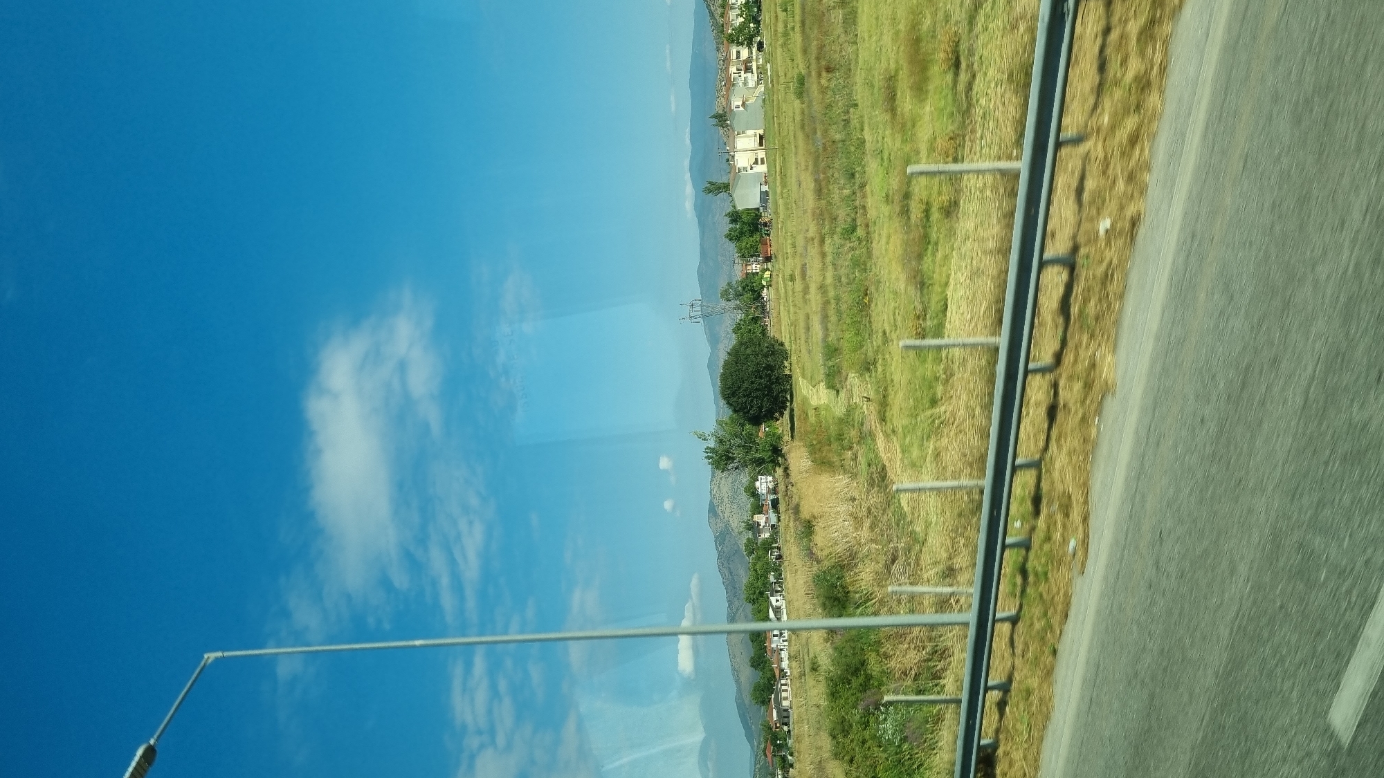 The image captures a rural landscape with a clear blue sky and scattered clouds. In the foreground, a gray metal guardrail runs horizontally across the bottom of the frame, with a road visible below it. Beyond the guardrail, a dry, grassy field stretches towards a treeline and a small settlement of white buildings. A tall, silver street lamp stands on the left side of the image. In the middle ground, a large, dark green, rounded tree is prominent, and a power line tower can be seen further in the distance. The background is dominated by rolling hills and mountains. The scene appears to be the outskirts of the town of Farkadona, Greece, suggesting a roadside view during daylight hours. There are no people or animals visible in the image.