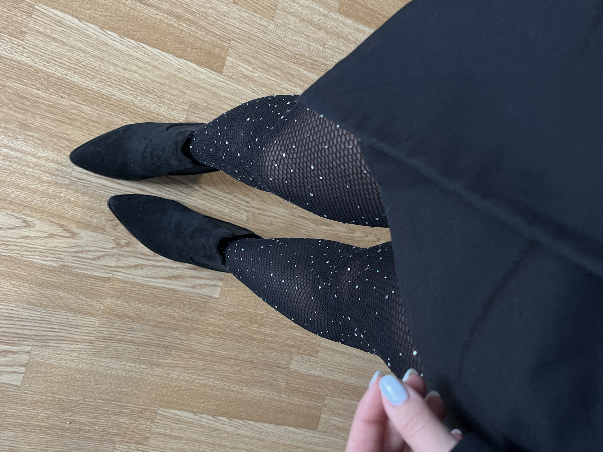 The image presents a top-down view of a person's lower body, showcasing a fashionable outfit against a light brown wooden floor. The individual is wearing black pointed-toe ankle boots, which appear to be made of a suede-like material. Their legs are adorned with black patterned tights featuring a fishnet-like texture, embellished with numerous small, sparkling elements, likely rhinestones or glitter, distributed across the fabric. A dark, solid black garment, possibly a skirt or dress, covers the upper thighs, partially obscuring the top portion of the tights.
In the lower right quadrant of the image, a hand with neatly manicured nails, painted a light pastel blue, is visible, gently resting near the leg or the dark garment. The wooden floor beneath has a distinct plank pattern, suggesting an indoor setting. The image focuses on the attire and provides no specific cues regarding the time of day or weather. No visible text is present in the image. The scene is located in Shushary, Russia.