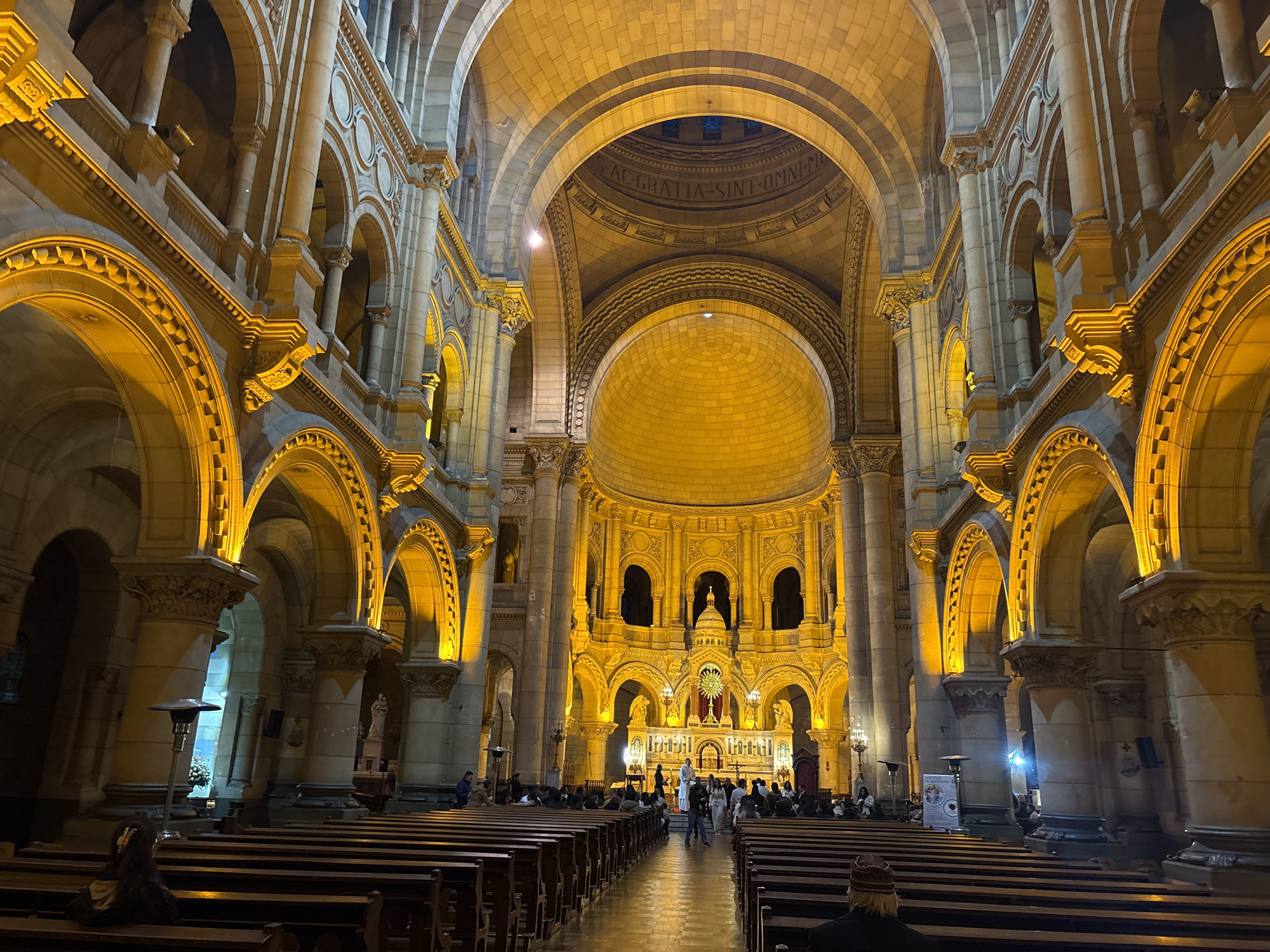 The image depicts the interior of the Basilica of Our Lady of Perpetual Help, located in Santiago, Chile. The grand hall of the church is illuminated by warm yellow lights, highlighting the intricate architectural details of the vaulted ceilings, arches, and columns. Wooden pews are arranged in rows, facing the altar at the far end of the hall, where a crucifix and religious imagery are visible. Several people are seated in the pews or walking through the aisle, suggesting a service or a visit by parishioners and tourists. The scene conveys a sense of solemnity and architectural grandeur characteristic of a large religious structure.