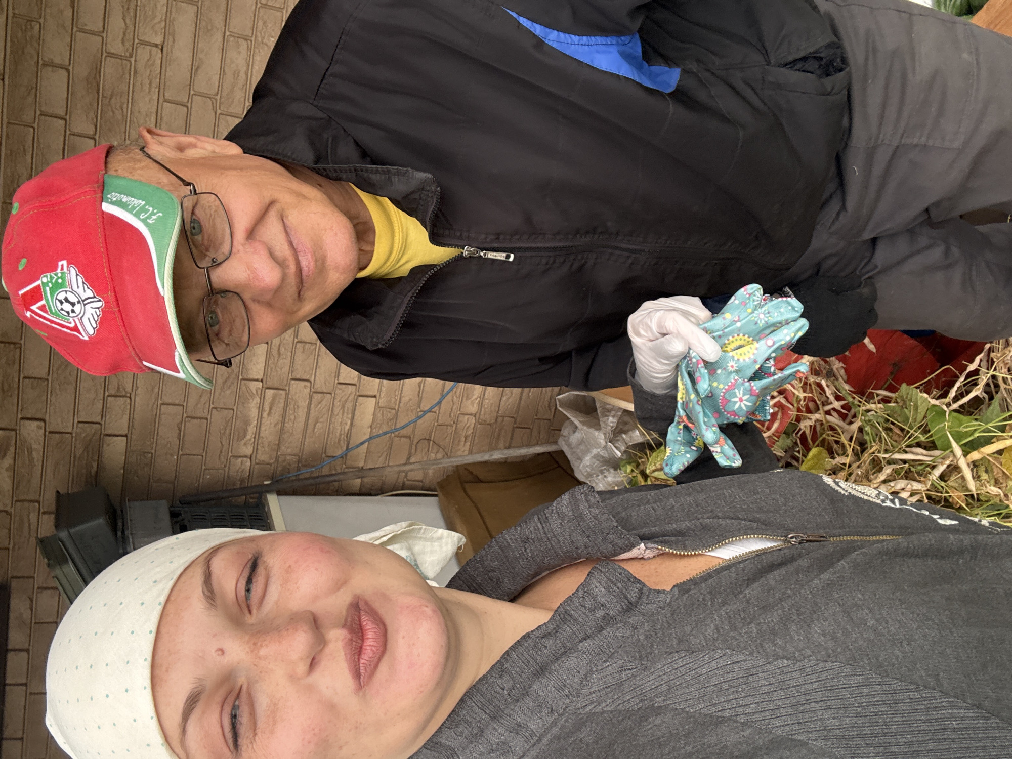 In Fominka, Russia, a woman and an older man pose for a selfie outdoors. The woman, in the foreground, wears a white headscarf and a grey zip-up jacket, her face turned towards the camera with her lips pursed. She has freckles and is wearing light makeup. The man, slightly behind her, sports a red baseball cap with a logo and a black jacket over a yellow shirt. He is wearing glasses and white gloves. He holds a pair of patterned gardening gloves in his gloved hand. Behind them, a brick wall and some outdoor storage containers are visible. The ground is covered with dry vegetation, suggesting a garden or yard setting.