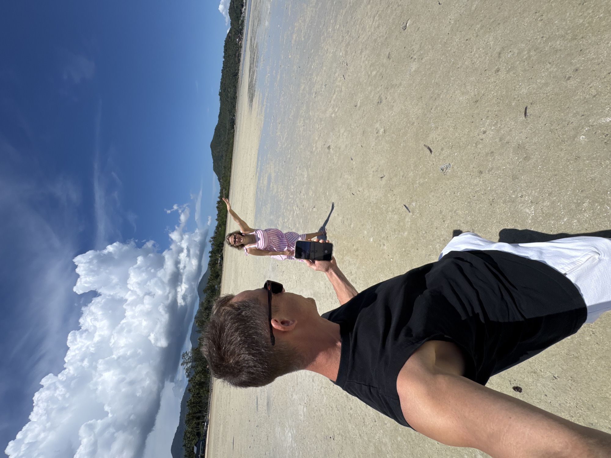 This is a selfie-style, eye-level, full-body shot of a man and a woman on a beach, likely on Koh Phangan, Thailand. The man is in the foreground, facing left and holding a phone, presumably to take the photo. He is wearing a black sleeveless shirt and white pants. The woman is in the mid-ground, facing the camera, wearing a pink and white striped dress and sunglasses. She is raising her left arm and pointing towards the distance. The beach is sandy and appears to be low tide, with shallow water covering parts of the sand. In the background, there are palm trees, vegetation, and hills, with a clear blue sky and scattered white clouds above. The scene is brightly lit by sunlight.
