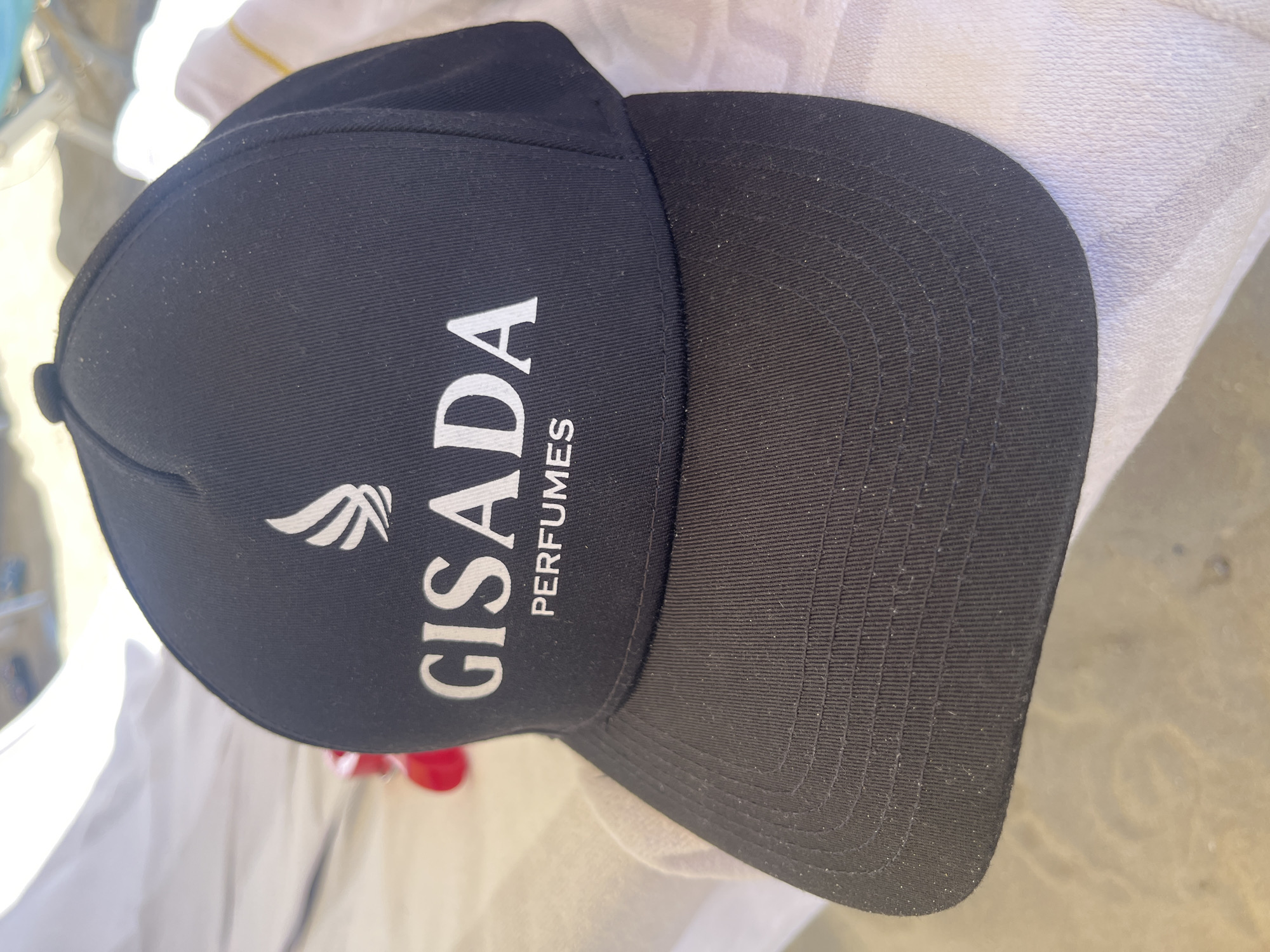 The image shows a black baseball cap with the logo "GISADA PERFUMES" in white on the front. The cap is sitting on a white, textured surface that looks like fabric. The location is unknown.
