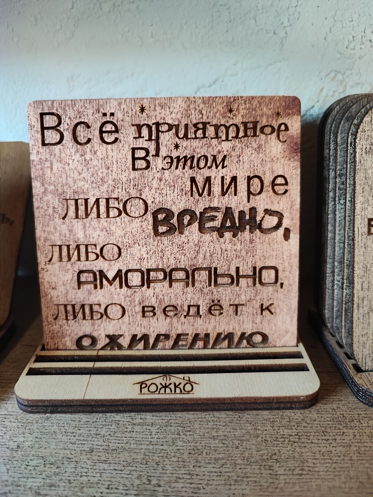 The image shows a wooden decorative plaque with Russian text carved into it. The plaque is displayed on a wooden stand. The text on the plaque appears to be a quote or saying. The plaque has a rustic, distressed wood finish. The stand is made of lighter colored wood and has grooves to hold the plaque upright. The background is a light-colored textured wall. The scene is indoors, likely in a shop or home. The bottom of the plaque has a logo that reads "РОЖКО". The context of Pyatigorsk, Russia is not evident from the image itself.