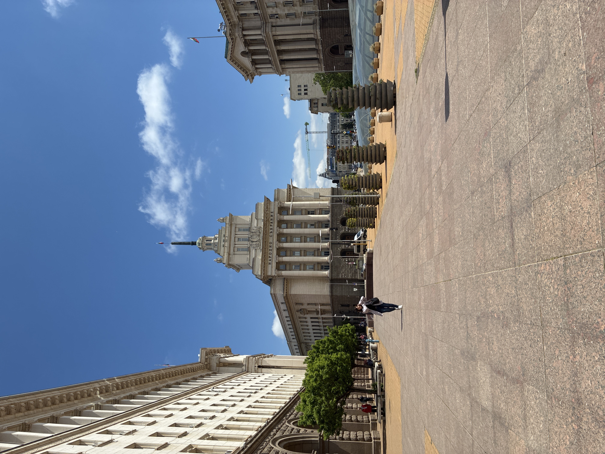 The image is taken at a ground-level perspective in Sofia, Bulgaria, featuring the city's architectural landscape under a partly cloudy sky. The most prominent building is the National Assembly of Bulgaria with its distinctive tower. Flanking the National Assembly building on either side are other large, symmetrical buildings.
The scene is a pedestrian plaza paved with red and beige stone tiles. Lining the plaza are stacked stone planters with greenery. A few individuals are walking along the plaza, including one person in a white coat, black pants, and white sneakers who appears to be talking on a cell phone. There are trees along the building on the left side of the plaza.
In the background, partially obscured by buildings and sky, a crane is visible. The overall ambiance suggests a bright, sunny day in an urban setting.