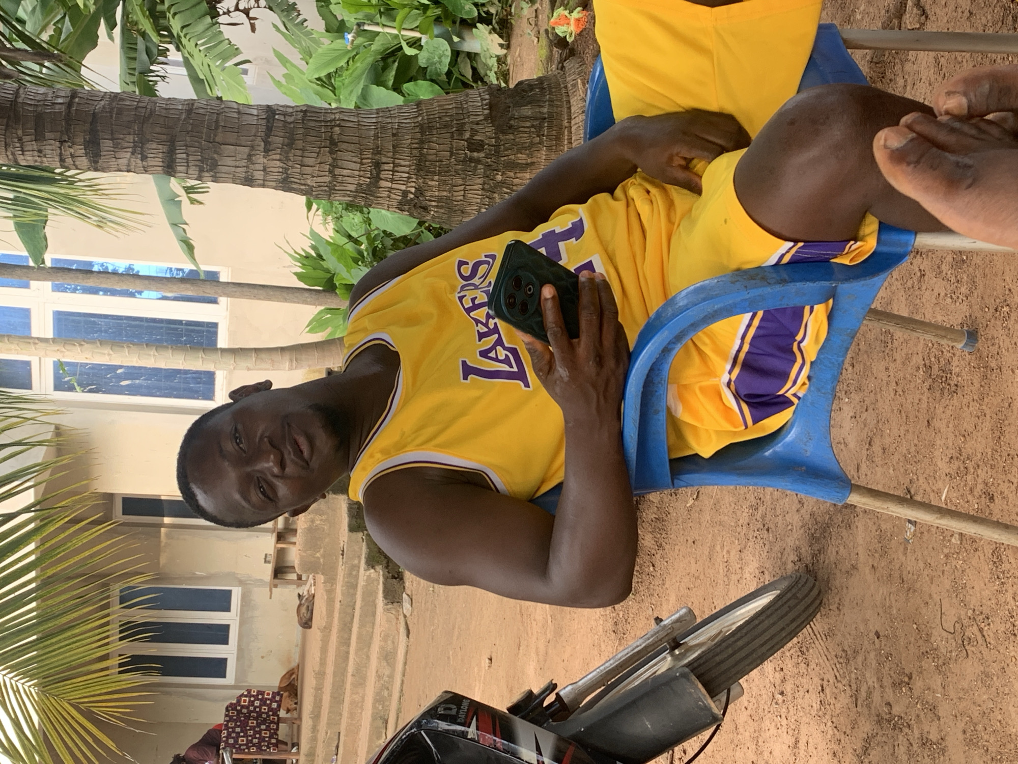 A Black man wearing a yellow Los Angeles Lakers basketball jersey and shorts sits in a blue plastic chair outdoors, holding a black smartphone in his right hand. He is positioned in the foreground, with a motorbike to his left and a palm tree to his right. Behind him, a building with large blue-tinted windows and a palm frond are visible. The ground is sandy and brown, with some loose gravel. In the background, two dogs lie on the steps of the building, and a person wearing a maroon shirt sits at a table covered with a patterned cloth. The scene appears to be in Akwanga, Nigeria, with the architecture and vegetation suggesting a warm, tropical climate.