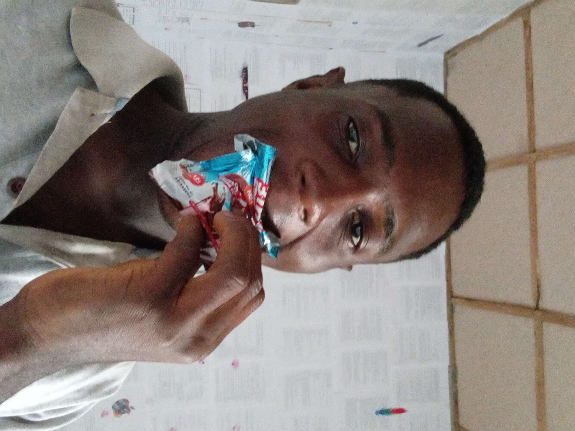A close-up shot shows a dark-skinned man, likely in his late teens or early twenties, holding the torn wrapper of a small candy or chocolate bar in his mouth. His right hand is holding the wrapper, suggesting he is in the process of opening the candy or has just finished eating it. He is wearing a light gray shirt with a button visible. The background features a wall covered in white wallpaper with faint markings or text. To the right, a section of the wall appears to be made of light-colored panels separated by wooden strips. The location and city are Akwete, Nigeria.