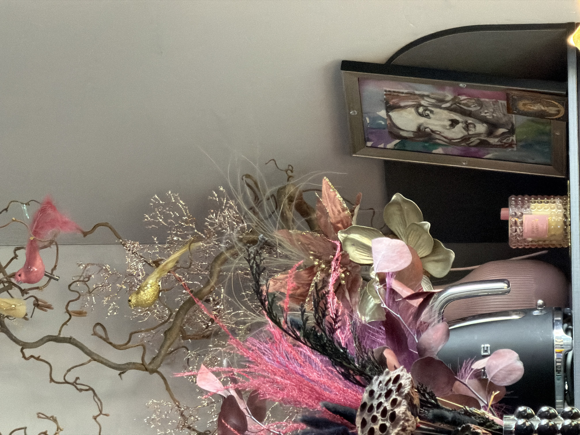 The image depicts an ornate, still-life arrangement. In the background, a framed artwork displays a face rendered in what appears to be a watercolor or mixed media style, with muted colors. Below the artwork, and towards the right, is a collection of objects on a shelf, including a perfume bottle, a round, pink item, and what appears to be a small, gray appliance.
The foreground is dominated by a complex arrangement of floral and decorative elements. Twigs, some adorned with small, artificial birds and shimmering gold accents, create a sprawling structure. Bright pink feathery plumes and gold-colored flowers are intermixed with darker foliage. A lotus pod is also visible near the bottom.
There is no information regarding the location as Moscow, Russia.