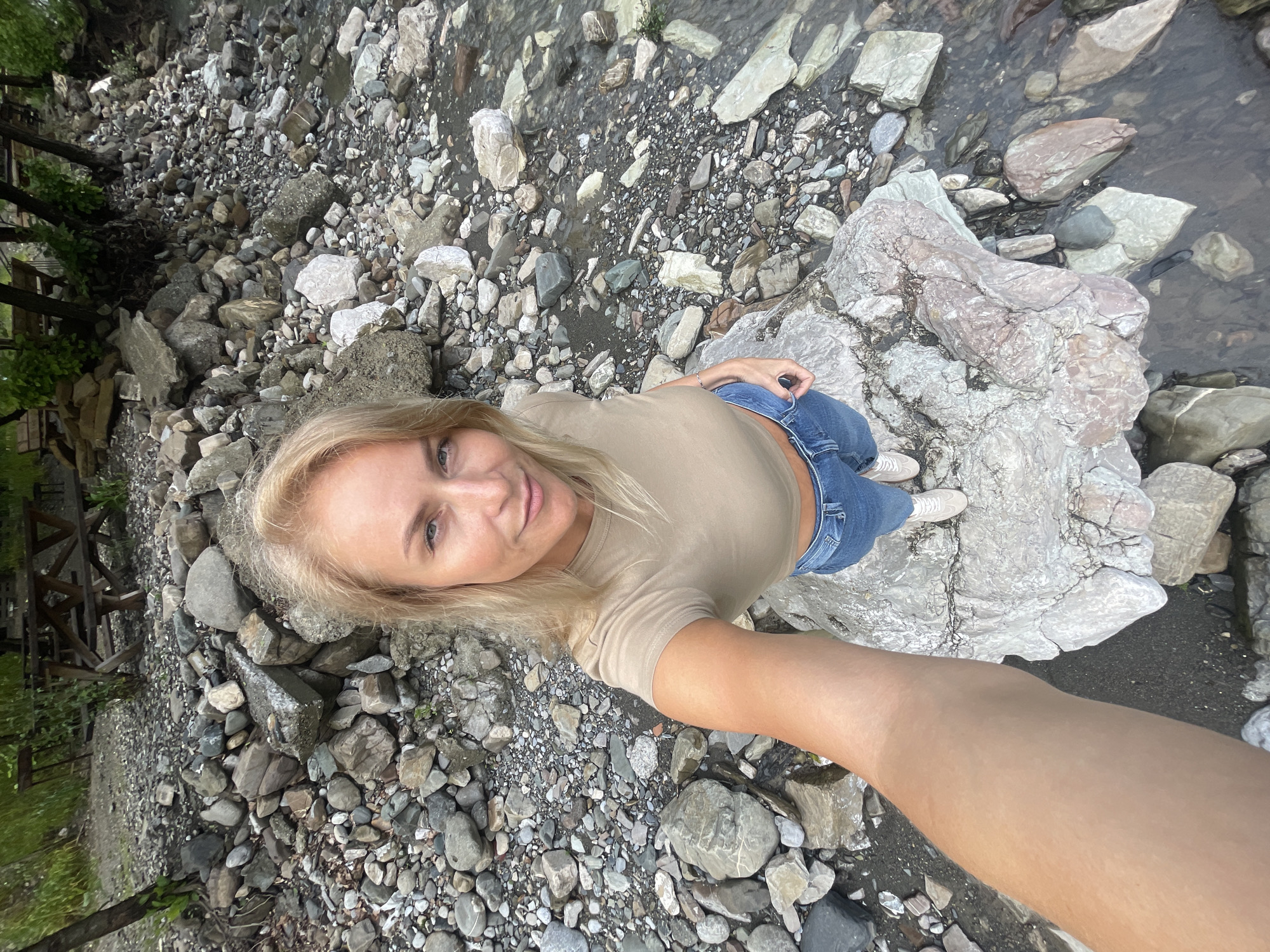 A blonde-haired woman stands on a large, light-colored rock amidst a rocky riverbed in what is likely the city of Sochi, Russia. The riverbed consists of various sizes and colors of rocks and pebbles. The woman is wearing a beige shirt, blue jeans, and white sneakers. She is taking a selfie, extending her arm towards the camera. A small stream of water flows nearby. In the background, a wooden structure is partially visible amidst greenery.