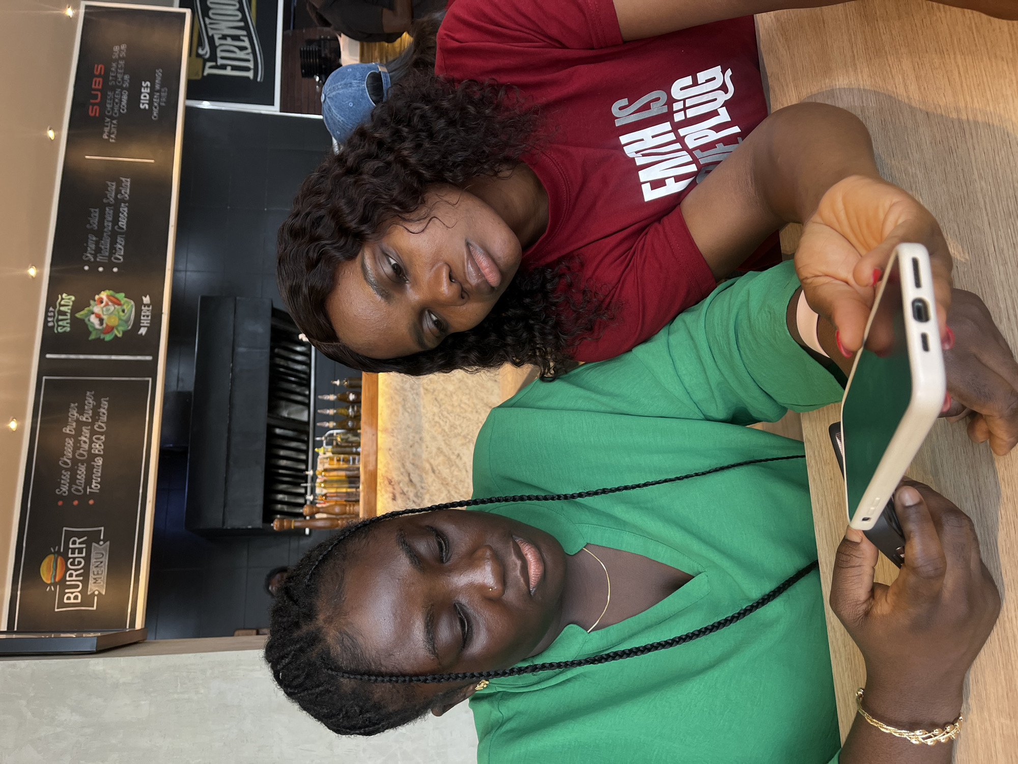 The image depicts two women seated at a light-colored wooden table within a casual restaurant environment, specifically identified as Firewood Restaurant in Abuja, Nigeria, based on signage visible in the background. Both individuals are focused on a smartphone held by the woman on the left.
The woman on the left has a dark complexion, wears her hair in thin braids, and has a small stud piercing on her left nostril and small gold-colored earrings. She is dressed in a long-sleeved V-neck top of a vibrant green color and a thin gold-colored chain necklace. On her left wrist, she wears a gold-colored braided bracelet. Her right hand holds a white-cased smartphone, orienting its screen downwards, while her left hand is also positioned near the device.
The woman on the right also has a dark complexion, with dark, wavy hair. She is wearing a red short-sleeved t-shirt with white text on the front, partially visible as "FAITH IS THE PLUG." Her right hand, featuring red nail polish on the fingernails, rests on the table. She is leaning slightly towards the woman on the left, directing her gaze downwards at the smartphone screen.
In the background, overhead black chalkboard menus display food options: "BURGER MENU" (featuring Swiss Cheese Burger, Classic Chicken Burger, Tornado BBQ Chicken), "BEST SALADS" (including Shrimp Salad, Mediterranean Salad, Chicken Caesar Salad), "SUBS" (listing Philly Cheese Steak Sub, Fajita Chicken Cheese Sub, Combo Sub), and "SIDES" (Chicken Wings, Fries). Behind the individuals, dark kitchen counters, an exhaust hood, and shelves with various bottles and shakers are visible. A person wearing a blue baseball cap is partially visible seated further back to the right, and another individual's arm is seen on the far right. The establishment's name, "FIREWOOD," is displayed in large lettering on a wall section to the right.