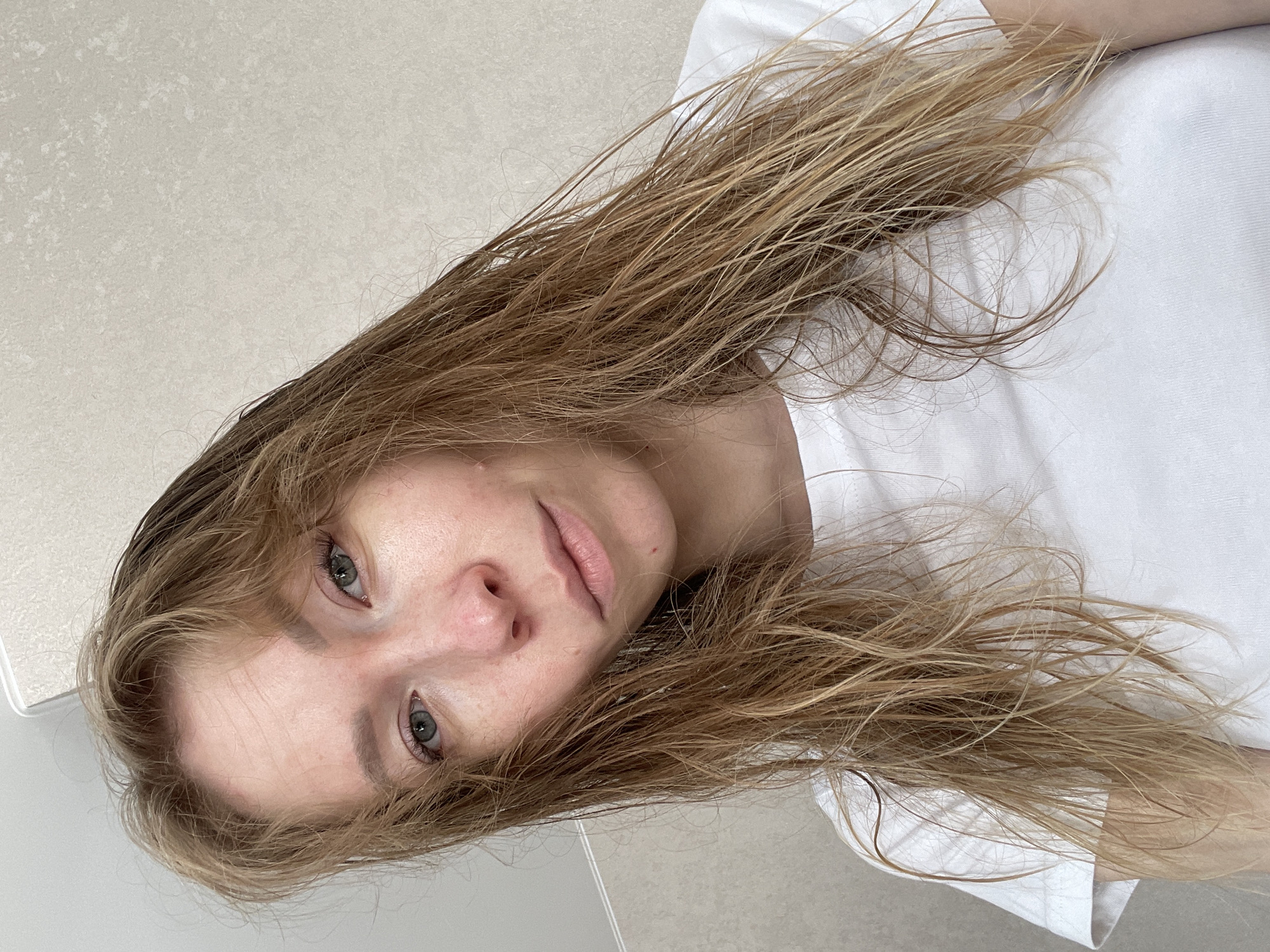 This medium close-up portrait, taken indoors in Perm, Russia, features a young woman with long, light brown hair that appears slightly damp or styled, cascading across the frame. Her face is central, oriented diagonally with her head towards the upper left, and she looks directly at the camera with a neutral, calm expression. She has blue-grey eyes and naturally colored lips; her skin shows some natural texture and a small blemish above her upper lip. She is wearing a simple white t-shirt. The background consists of light-colored, textured tiles, with a visible grout line in the lower right, suggesting an indoor setting like a bathroom or a tiled room. The lighting is soft and even. There is no visible text.