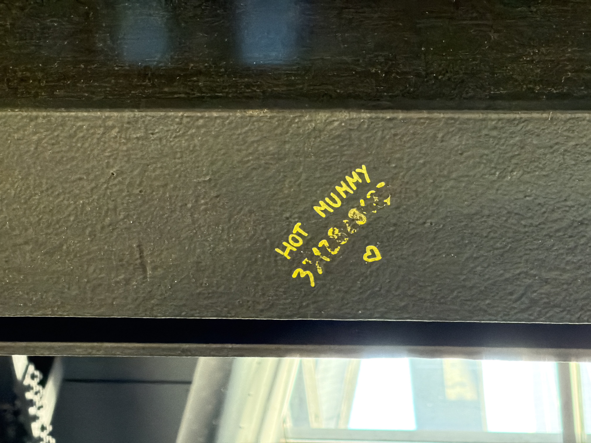 A close-up shot shows graffiti in yellow marker on a dark, textured surface. The graffiti reads "HOT MUMMY 3/12/2023" followed by a heart symbol. The background suggests the edge of a window frame, possibly in an interior setting. The location is not discernible from the image.