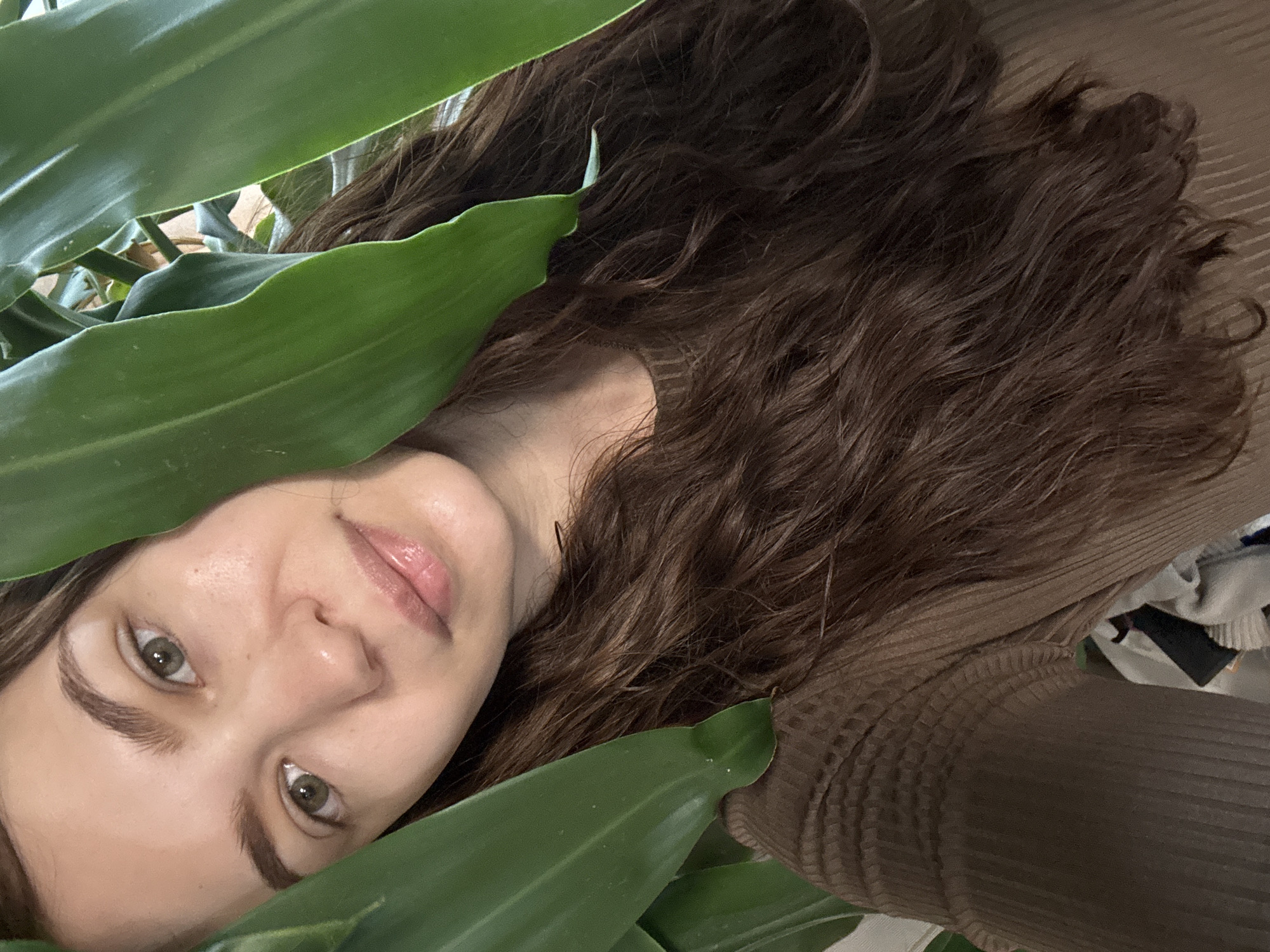A woman with brown hair is partially obscured by green leaves. She has light skin, green eyes, and pink lips. She is wearing a brown ribbed top. The background is unclear but appears to be a cluttered indoor space. There is no indication of the location being Nizhnyaya Gar', Russia.