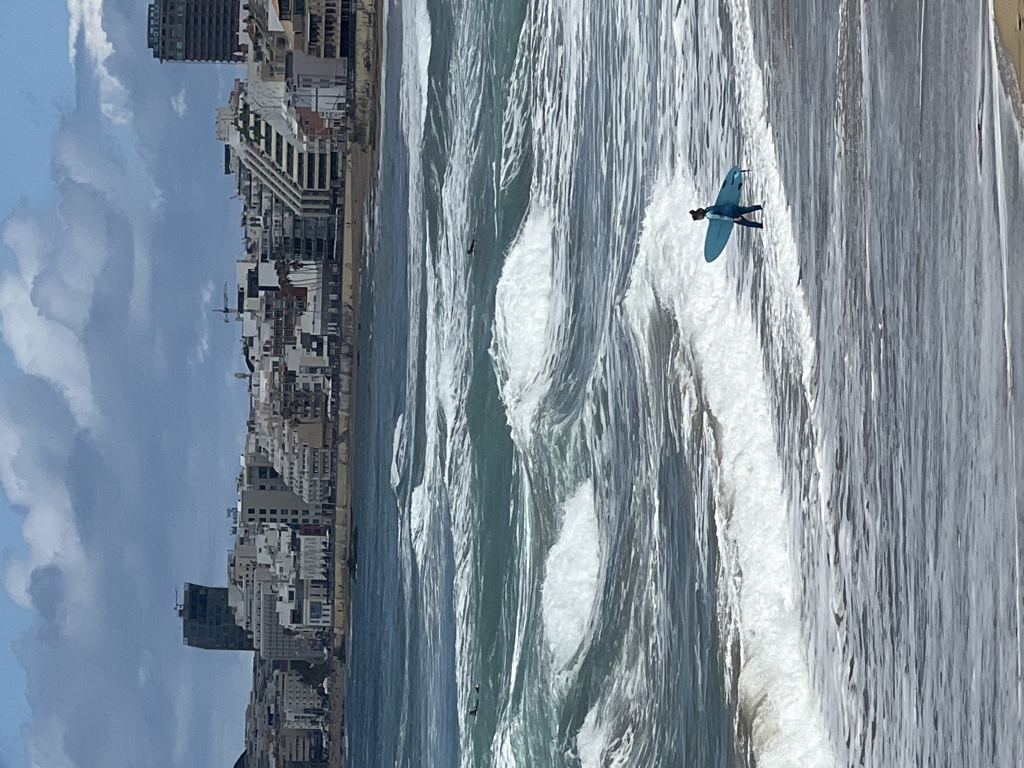 This image captures a vibrant coastal scene in Las Palmas De Gran Canaria, Spain, on a bright, partly cloudy day. The main subject is a surfer riding a light blue surfboard on a breaking wave in the Atlantic Ocean. The surfer, dressed in a dark wetsuit, is positioned towards the right side of the frame, actively engaging with the ocean swell. Another dark, smaller figure, likely another surfer, can be seen further out in the water amidst the waves.
The ocean dominates the middle and foreground, displaying multiple parallel waves with foamy white crests against varying shades of blue and green water. Along the left side of the frame, stretching horizontally, is the dense skyline of a coastal city. Numerous multi-story buildings, predominantly light-colored, line the shore. A tall, darker building stands out among them, and faint text resembling "HOTEL" is partially visible on its upper facade. A construction crane is also discernible amidst the urban structures. A narrow strip of sandy beach separates the city buildings from the ocean. The sky above the city is a pale blue with scattered fluffy white clouds.