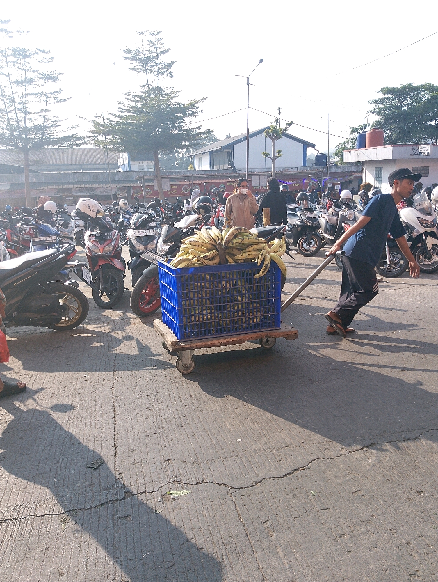 The image depicts a bright, outdoor scene in what appears to be a parking area or market in Lembang, Indonesia, judging by the presence of parked motorcycles and the architectural style of the buildings in the background. The sky is bright and overcast, casting soft shadows on the concrete ground, which is cracked and textured.
In the foreground, a man wearing a dark blue t-shirt, dark pants with white stripes on the side, and a black baseball cap is pushing a cart. The cart has a blue plastic crate filled with bananas, and it is positioned on a metal frame with small wheels. The man is actively engaged in moving the cart, with his right hand on the handle and his body leaning forward. He is wearing sandals.
To the left of the man and his cart, several motorcycles are parked. One black motorcycle is particularly visible in the mid-ground. Further in the background, more motorcycles are parked in rows. There are also people visible in the background, some standing near the motorcycles and others walking. One person in the mid-ground, to the left of the cart, is wearing a light brown jacket and a mask. Another person is wearing a dark shirt and a black helmet.
The buildings in the background appear to be low-rise structures, possibly shops or market stalls, with corrugated metal roofs and some painted walls. The overall impression is of a busy, everyday scene in a local Indonesian community.