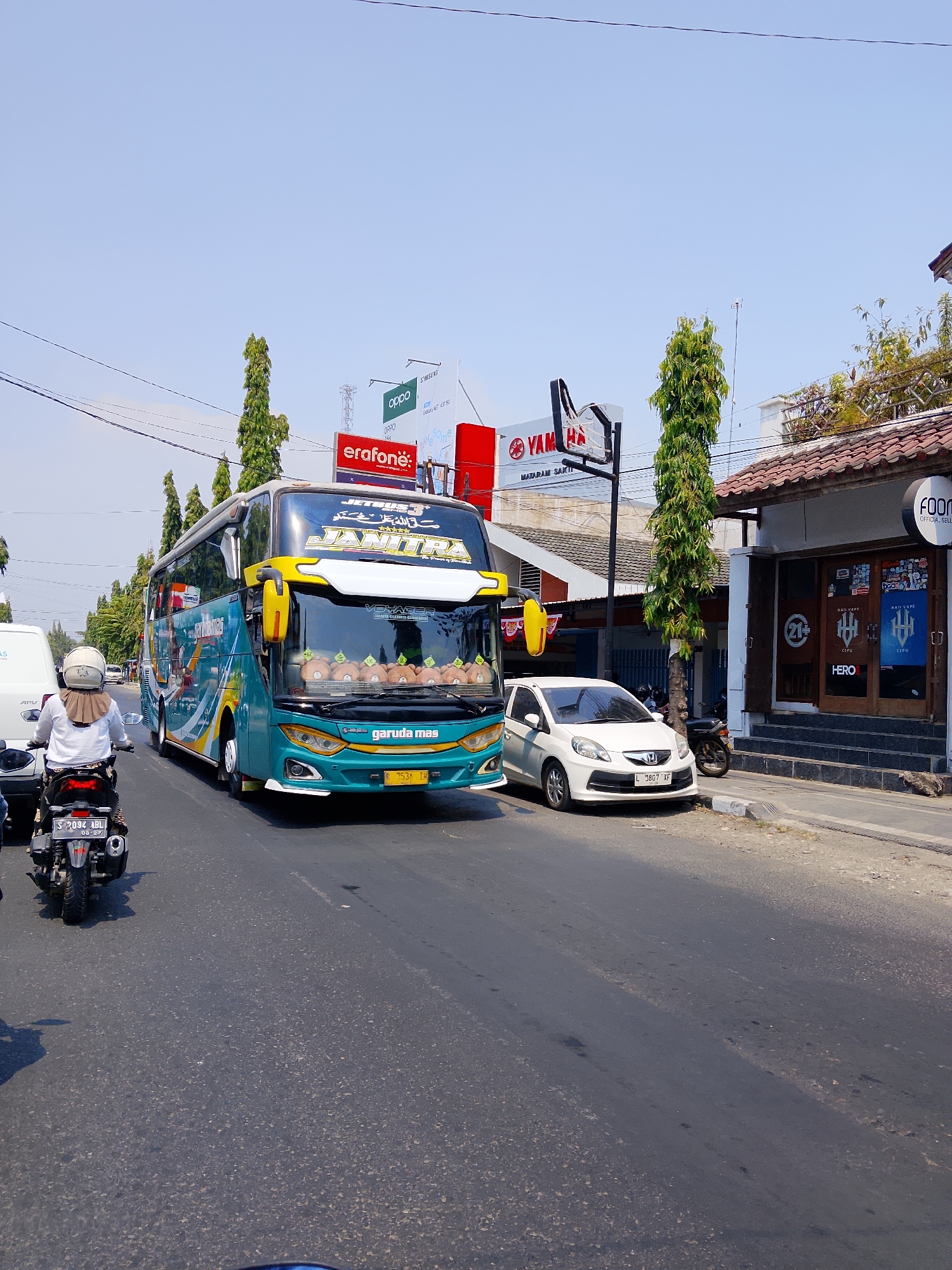The image captures a street scene in Ngelo, Indonesia, on a bright, sunny day. A large teal and yellow intercity bus with "GARUDA MAS" branding is the prominent subject, traveling on a paved road. The bus features "JETBUS" and "JANITRA" text on its front. In front of the bus, a white hatchback car is parked on the side of the road. To the left of the bus, a person on a black motorcycle with license plate "S 2034 ABL" is visible, moving in the same direction as the bus.
Along the right side of the road, a building with a tiled roof and large glass windows is present. Signs for "FOOD", "HERO", and "VAPE" are visible on the building's facade. Overhead, utility wires crisscross the sky. Behind the bus and the building, several tall, slender trees with dense green foliage line the street. Billboards advertising "OPPO", "SAMSUNG", and "YAMAHA" are also visible in the background, indicating commercial activity in the area. The sky is clear and blue, suggesting good weather conditions. The overall scene depicts a typical urban transportation environment in Indonesia.
