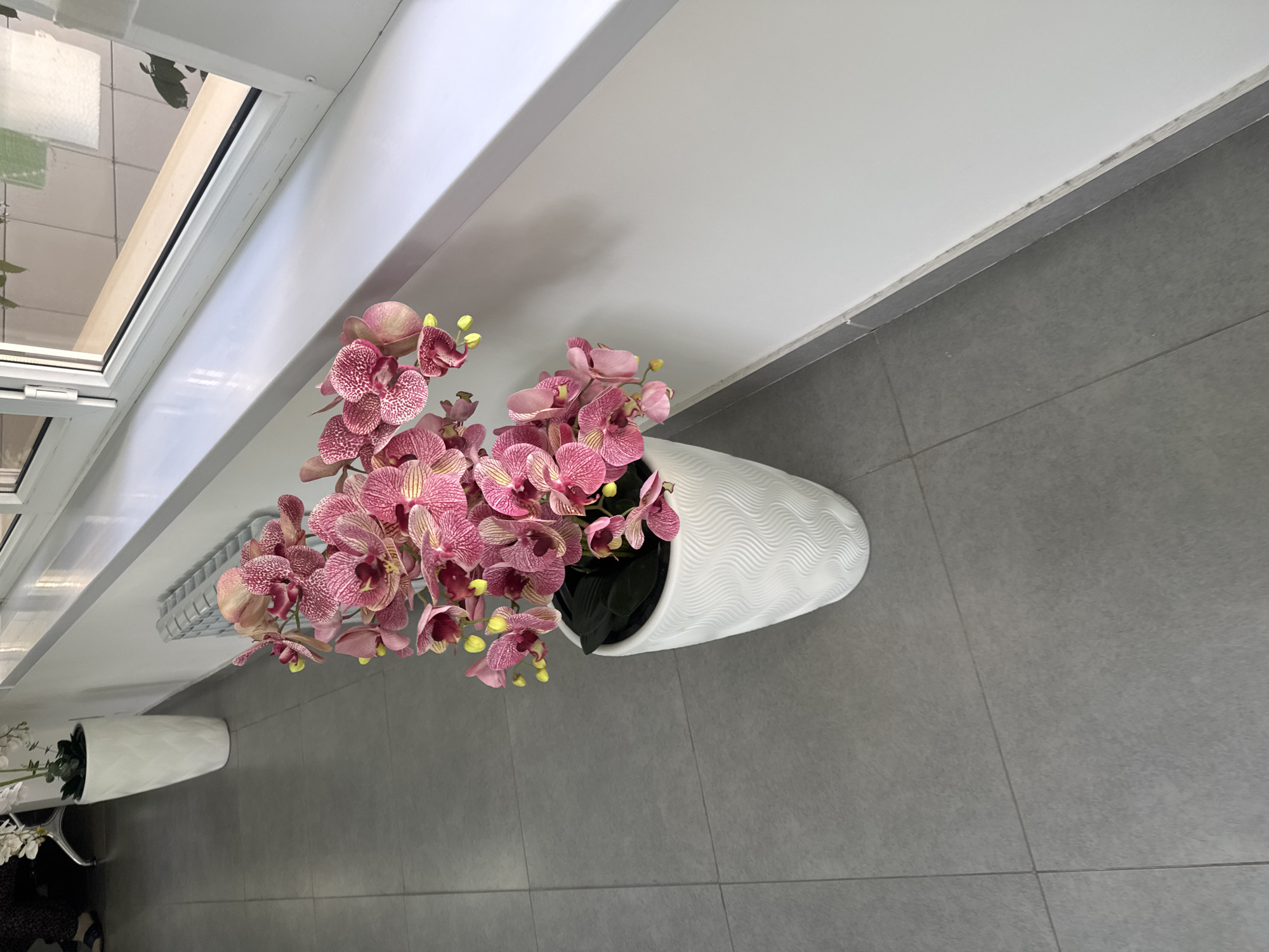 A high-angle medium shot shows a large, white, textured vase filled with artificial pink orchids. The vase sits on a gray tiled floor, positioned to the right of the frame. Behind the vase, a light-colored wall runs horizontally, with a window above it. To the far left of the frame, another tall white vase with a green plant is partially visible. The overall setting appears to be an indoor space, possibly an office or hallway, with a tiled floor and white walls. The lighting is bright and even. There are no people or animals visible in the scene. The location is specified as Shymkent, Kazakhstan.
