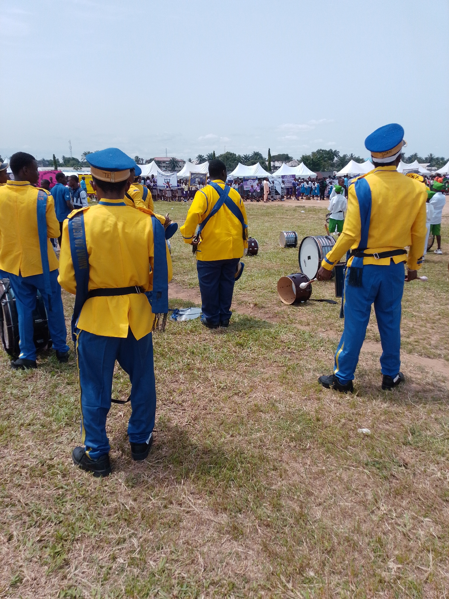 The image depicts a group of uniformed drummers standing on a grassy field in what appears to be Abak, Nigeria. They are wearing yellow jackets with blue accents and blue pants. In the background, there are tents and a crowd of people. Several drums are scattered on the ground.