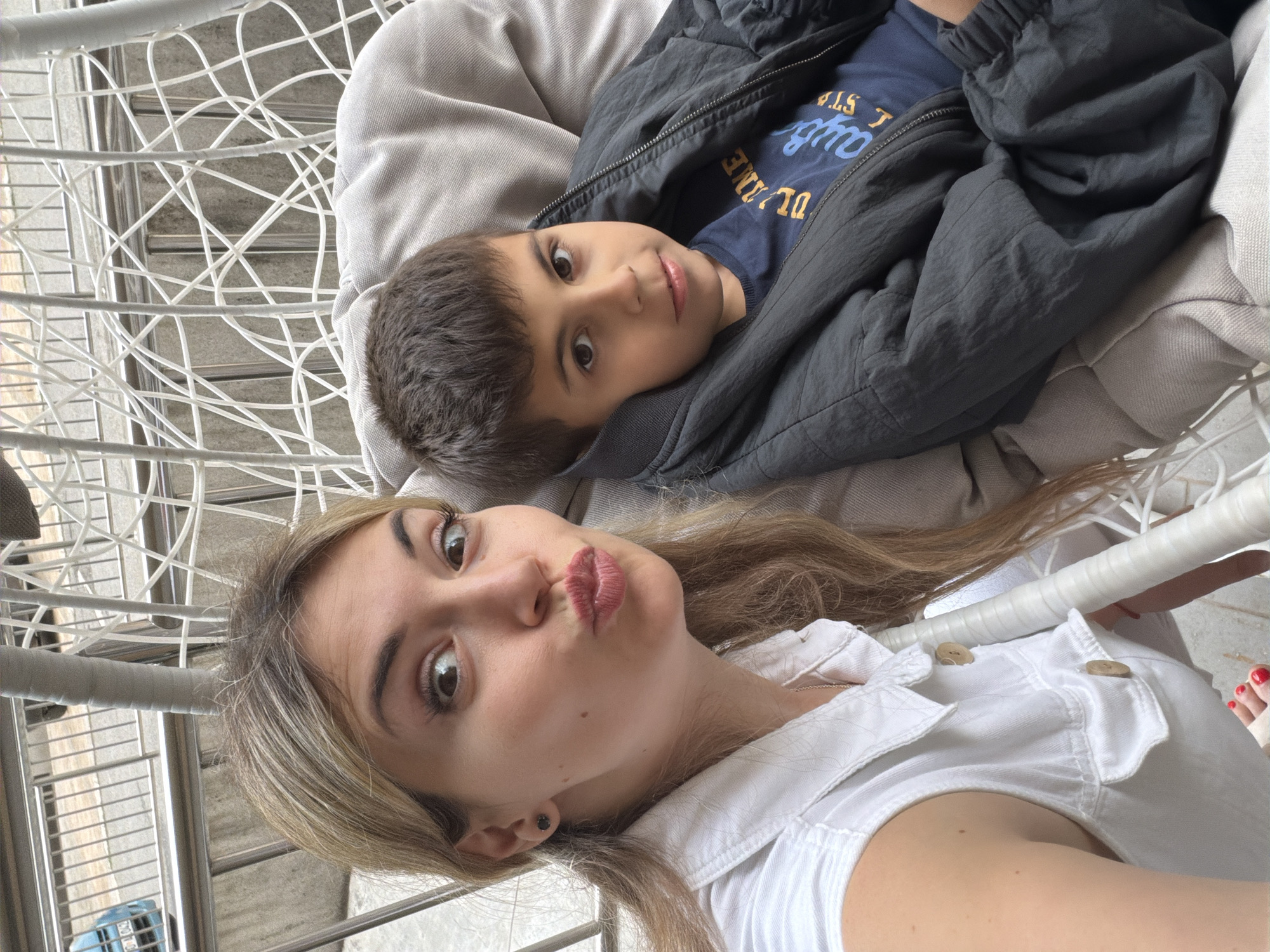 An upward-facing photo captures a woman and a child lying back, likely in a swing chair or similar outdoor furniture, taken from above. The image is rotated so the woman's head is at the bottom and the child's head is towards the top.
The woman, with long light brown hair and prominent eyebrows, has her lips pursed in a playful "kissing" gesture and looks directly at the camera with wide, dark eyes. She is wearing a white collared top or dress with visible wooden buttons. A dark earring is visible in her left ear, and her toenails, visible in one of the crops, are painted red.
Above her, a young boy with short dark hair is also looking towards the camera with wide, curious eyes and a slight, gentle expression. He is wearing a dark blue t-shirt with some yellow text on it, partially visible as "D L I N E", "S L S", and "q o", under a dark grey or black jacket.
Both individuals are resting on light grey cushions. The background reveals a white, woven or netted structure, characteristic of a hanging chair or a cocoon swing. Beyond this, there are glimpses of horizontal metal railings and a lighter-colored, possibly concrete, surface, suggesting an outdoor setting like a balcony or patio in Budva, Montenegro. The scene is brightly lit, indicating daytime.