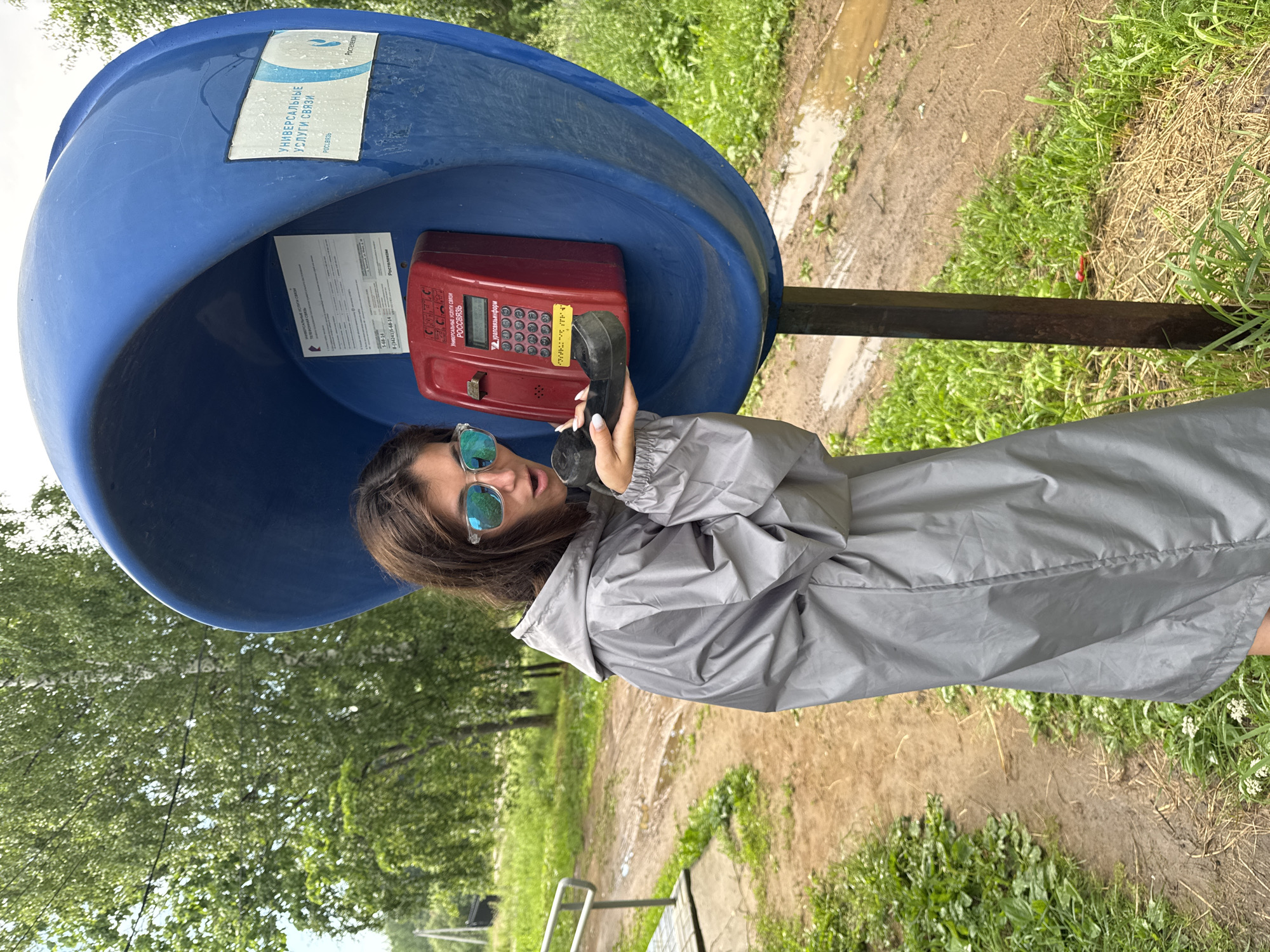 A young woman with dark hair and sunglasses stands in front of a blue, open-faced public telephone booth. She is wearing a grey hooded raincoat and holding a black phone receiver to her ear. The phone itself is red and mounted inside the booth. The background depicts a rural outdoor setting with green trees, a dirt path with puddles, and lush grass. A metal railing is visible to the left of the woman. The scene appears to be in the village of Vasyata, Russia, as indicated by the text on the phone booth.