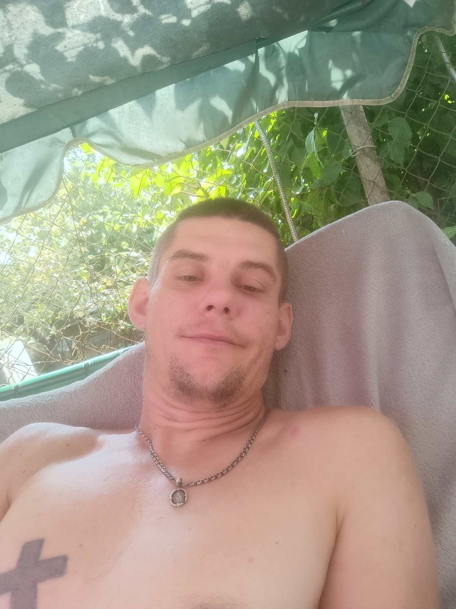 A shirtless man with a short buzz cut, a goatee, and a tattoo on his chest is reclining on a towel under a green canopy. He is wearing a silver chain with a circular pendant. The background shows a chain-link fence, green foliage, and sunlight filtering through. The man is looking towards the camera with a relaxed expression. The location is described as Zaliznyi, Russia.