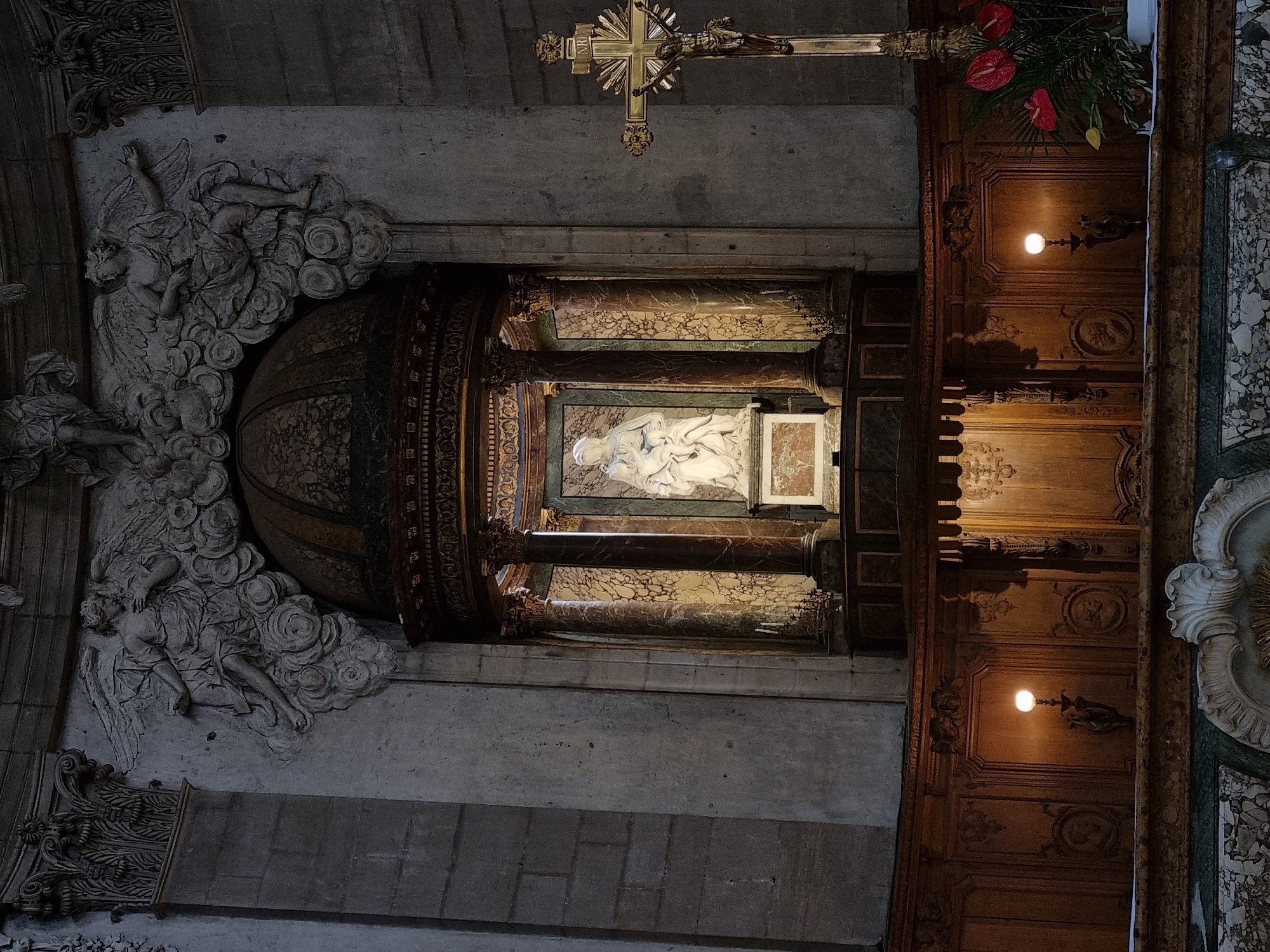 This image shows an interior view of a church, potentially located in an unknown city, likely in Europe. The scene is dominated by architectural and religious elements. A gold crucifix hangs at the top of the frame, while below, a complex altar or shrine is visible. This structure is adorned with ornate carvings, including marble, wood, and a relief sculpture depicting what appears to be figures reclining on clouds. The altar is also embellished with flowers and two lit sconces. The environment is dim, with the lighting primarily coming from the sconces and possibly other unseen sources, highlighting the details of the architecture and sculptures.