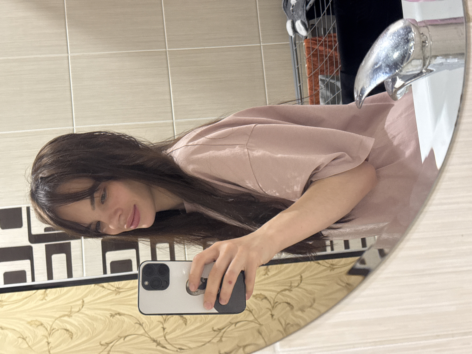 The original image is rotated 90 degrees clockwise, displaying a mirror selfie of a young woman with long, dark hair, wearing a light pink t-shirt. She is reflected in a round mirror, holding a white smartphone in her left hand. The smartphone features three visible camera lenses on its back and a black pop socket/grip. The woman is smiling gently, looking towards the camera.
The scene, located in Rostov-on-Don, Russia, appears to be a bathroom or a similar interior space. Visible in the reflection are light beige square tiles on the upper wall. Below these tiles, a section with a black and white geometric pattern is present, followed by a golden-patterned wallpaper.
A silver-colored faucet is seen extending over a white sink basin in the reflection. Behind the faucet, a metal grid rack or shelf holding various items is partially visible. The lighting suggests a well-lit indoor environment. There is no visible text in the image.