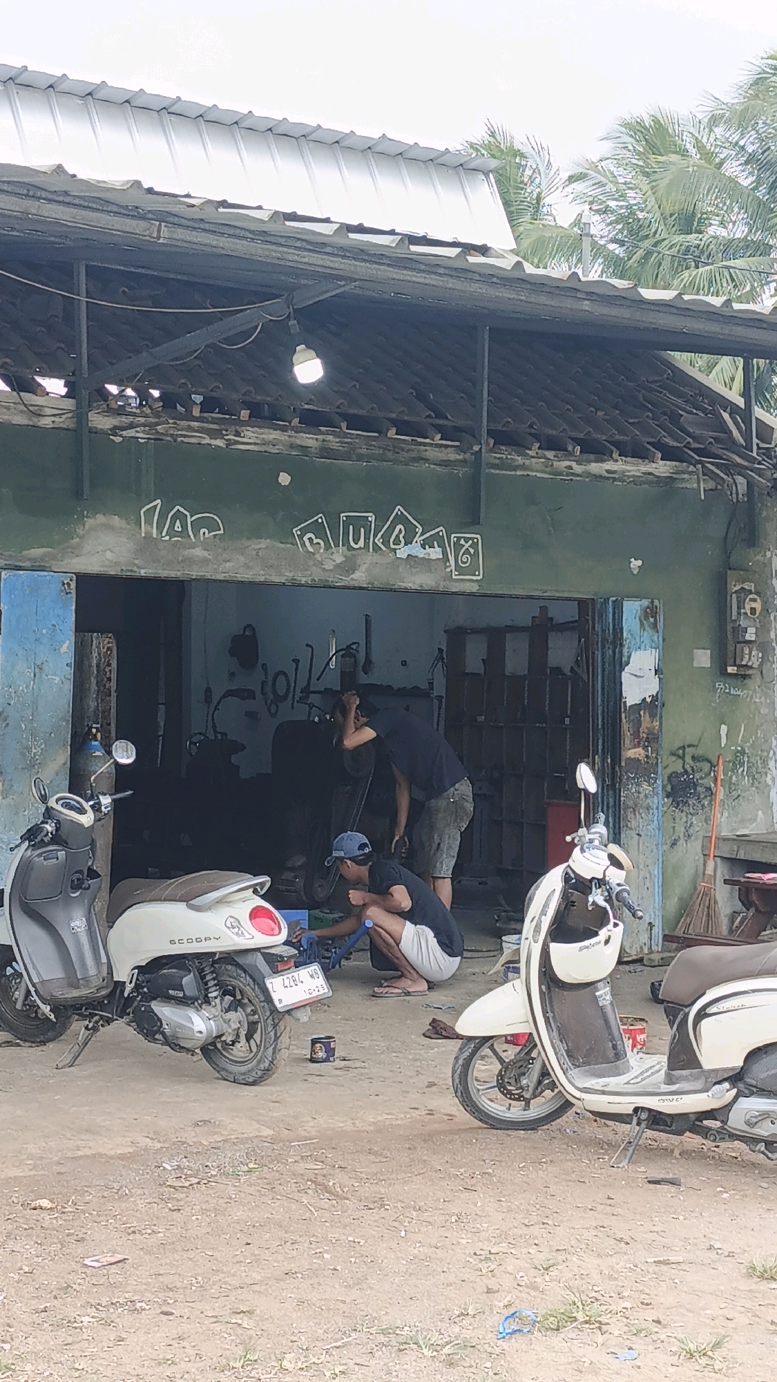 In Cibenda, Indonesia, two men are observed in front of a rundown workshop. The workshop has an open front with a corrugated metal roof supported by exposed beams. The building's exterior is painted a faded green, adorned with graffiti that appears to spell out "JAG MUA". Inside the workshop, a person is bent over, seemingly working on a motorcycle, while another person stands nearby, observing or assisting.
Two scooters are parked outside the workshop. The scooter on the left is light gray with a brown seat, and the scooter on the right is cream-colored with a brown seat. The ground in front of the workshop is dirt and gravel. A broom leans against the right side of the building, and a gas cylinder is visible near the left scooter. The overall environment suggests a utilitarian setting, possibly a motorcycle repair shop or a similar service establishment.