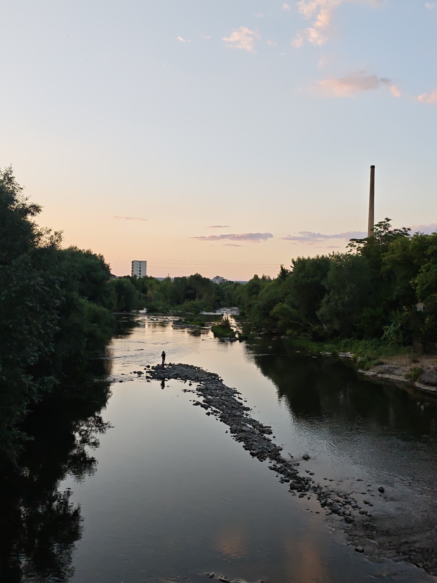 The image depicts a wide, calm river flowing through a verdant landscape, likely within the city of Lovech, Bulgaria, given the distinctive chimney in the background. The time of day appears to be dusk or dawn, with a soft, pastel-colored sky transitioning from pale blue to peachy hues.
In the mid-ground, a lone figure, silhouetted against the water, stands on a small, rocky island in the middle of the river. The figure's posture suggests they might be fishing or observing the surroundings. The riverbanks are densely lined with lush green trees, creating a natural, almost wild atmosphere despite the presence of distant urban structures.
To the right of the frame, a tall, slender brick chimney rises above the trees, a prominent industrial landmark characteristic of Lovech. In the distance, on the left side of the river, a modern, multi-story building stands out against the horizon, indicating the urban edge of the city. The water itself reflects the muted colors of the sky and the surrounding foliage, creating a serene and somewhat contemplative mood. The composition emphasizes the interplay between nature and the built environment, with the river acting as a central element connecting them.