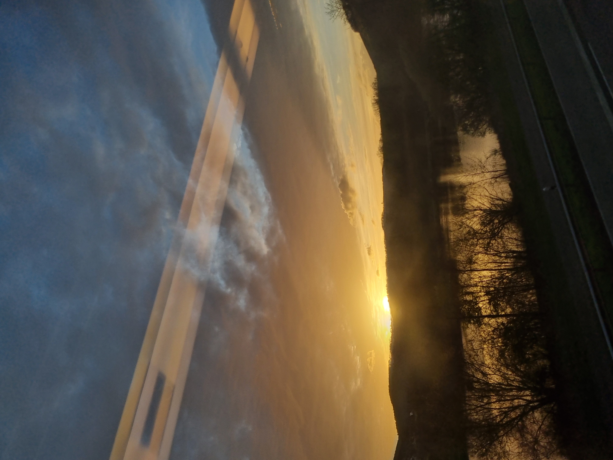 The image shows a reflection of a sunset and clouds in a window, likely in Saarburg, Germany. The reflection shows a golden sunset illuminating the sky and clouds, along with a silhouette of the Saarburg landscape, including a river, trees, and what appears to be a road. The presence of the reflection indicates the photo was taken indoors, perhaps from a building overlooking the landscape.