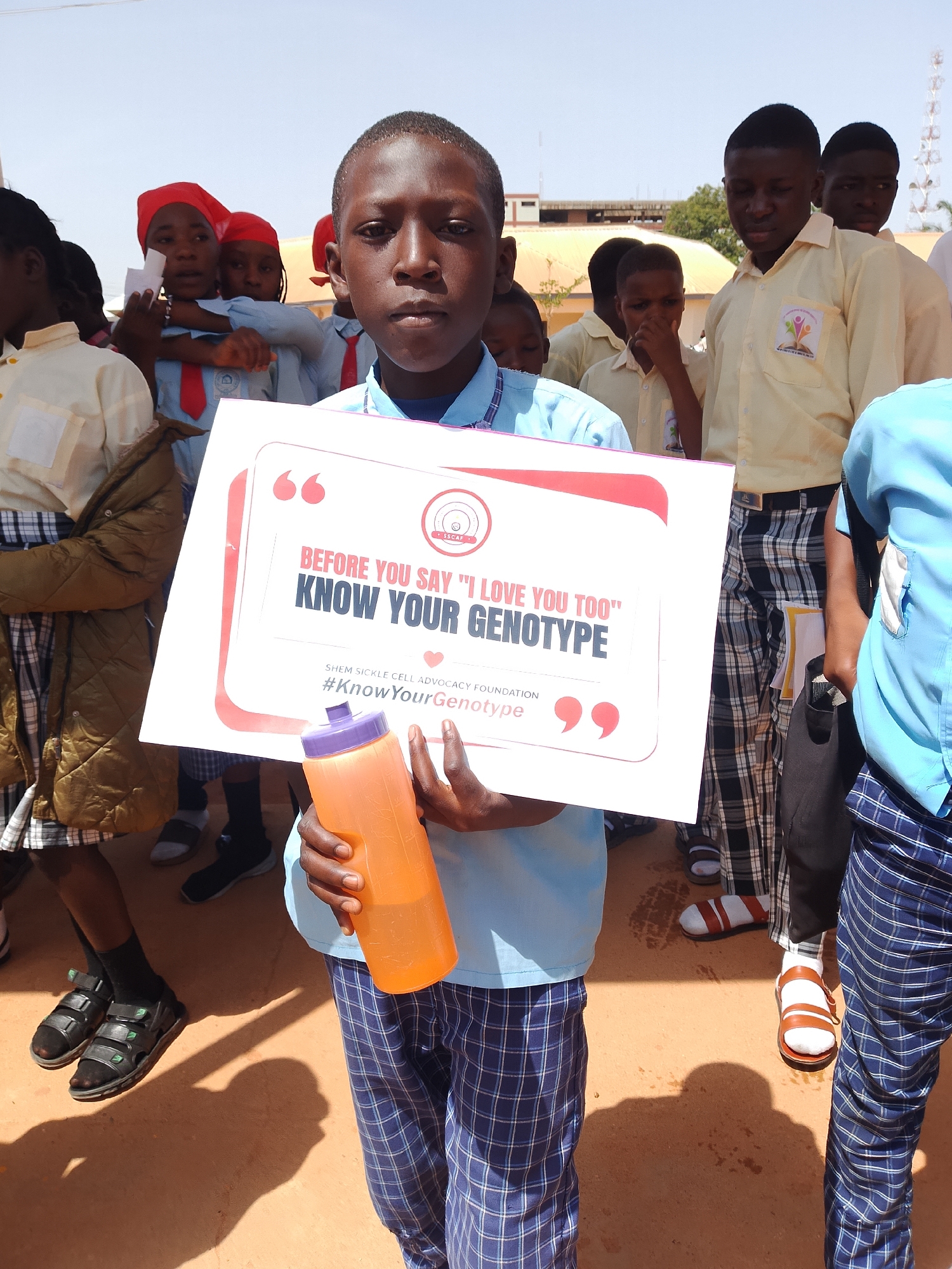 In a daylight outdoor scene in Jos, Nigeria, a group of young people, primarily children and teenagers, are gathered, possibly for an event or educational activity. The focus is on a young boy in the foreground, holding a sign that reads, "BEFORE YOU SAY 'I LOVE YOU TOO' KNOW YOUR GENOTYPE." The sign also features the logo of "SHEM SICKLE CELL ADVOCACY FOUNDATION" and the hashtag "#KnowYourGenotype." Several other individuals are visible behind him, some wearing light blue shirts and plaid pants, and others in yellow shirts. One person is holding an orange water bottle. The environment appears to be an open area with a building and trees visible in the background under a clear sky. The ground is a reddish-brown, suggestive of dusty terrain common in Jos. The overall atmosphere suggests a public health awareness campaign or a school-related event concerning sickle cell advocacy.