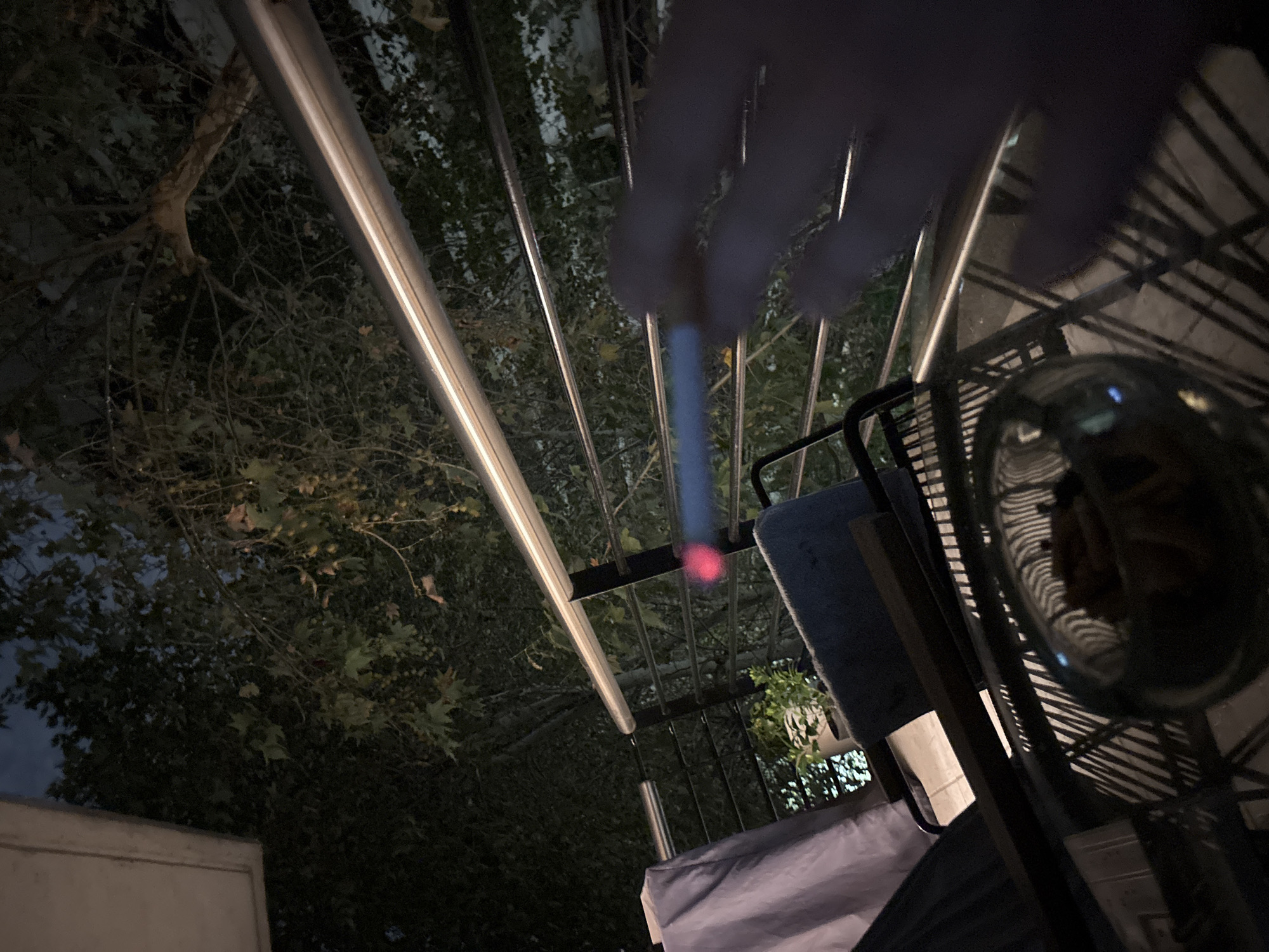 A nighttime, low-angle shot shows a partially obscured hand holding a lit cigarette. The background contains trees and a metal structure, possibly a pergola. Visible items include a metal chair and a glass. It is not possible to ascertain the location in Buenos Aires, Argentina.