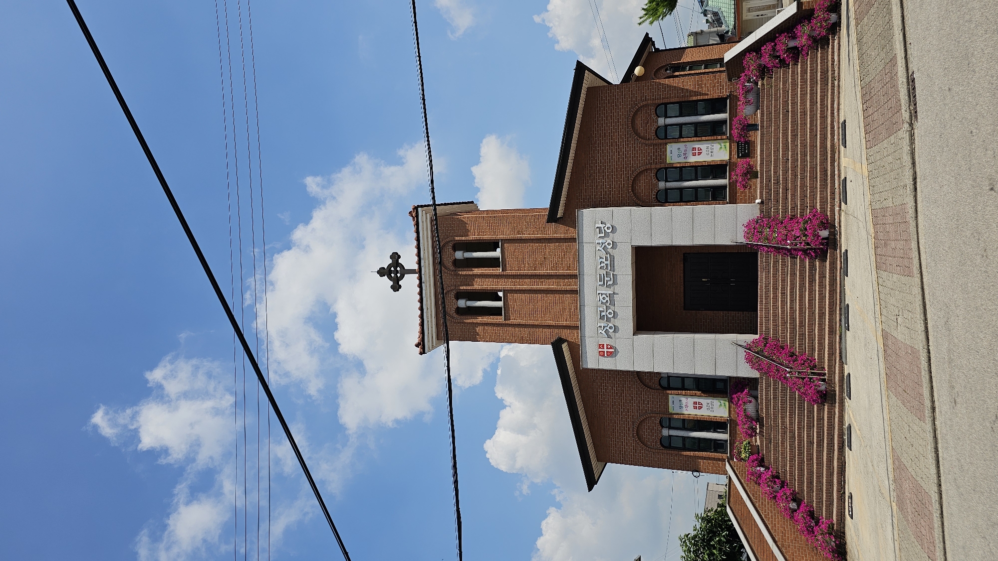 The image shows a brick church with a bell tower topped by a cross, located in Asan-si, South Korea. The church has a prominent set of stairs leading to the entrance, decorated with numerous pink flowers. The building features arched windows and a sign above the entrance with Korean characters and a cross emblem. The sky is blue with white clouds, and power lines are visible crossing the upper part of the frame.