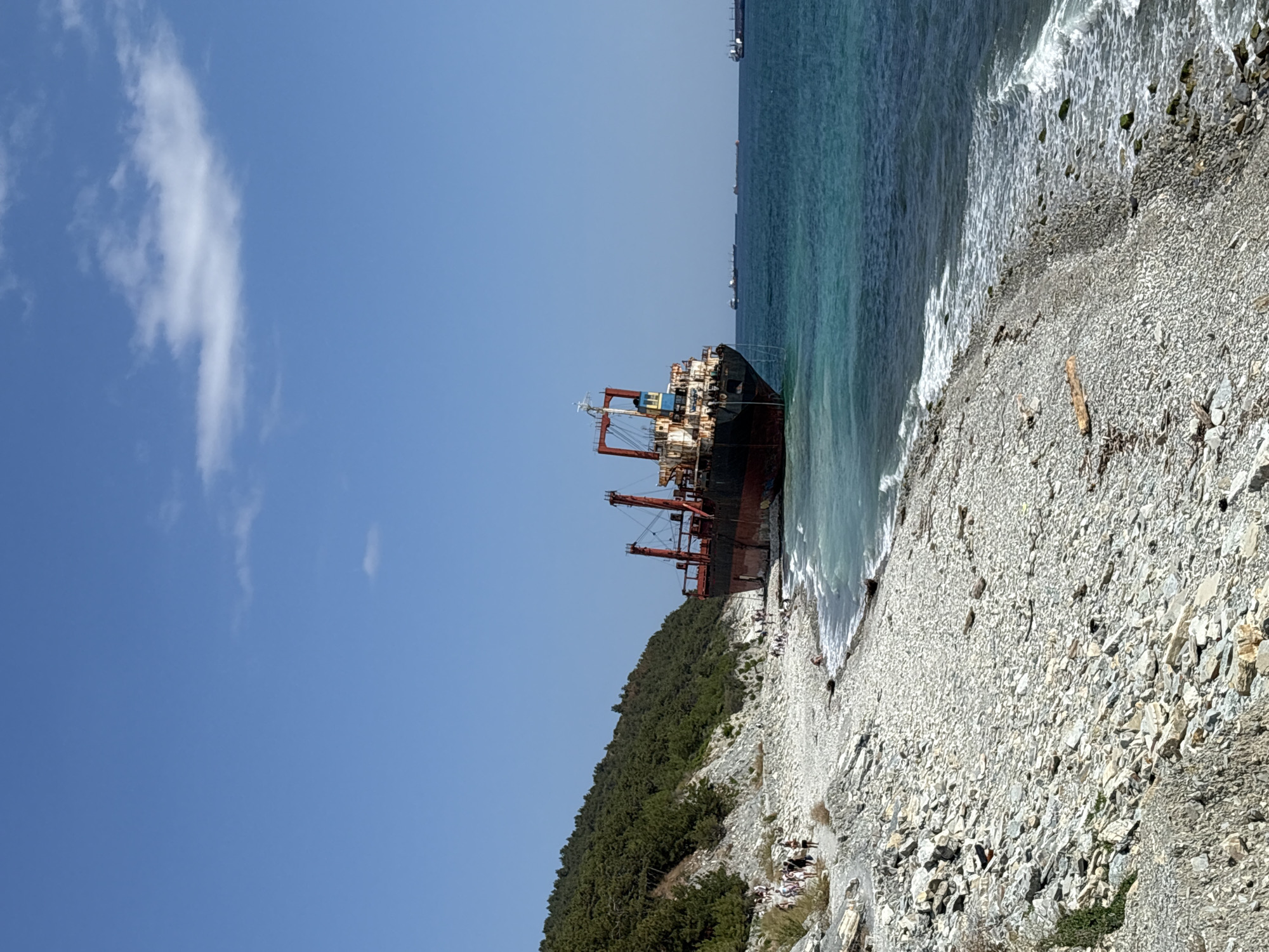 The image captures a view of a rocky coastline in Kabardinka, Russia, with a beached cargo ship dominating the scene. The ship, a large vessel with a red hull and superstructure, is grounded on the shore, its stern facing the camera. Its decks are weathered, suggesting it has been there for some time.
The shoreline is covered in a multitude of light-colored, smooth pebbles. A path runs along the edge of the beach, where several individuals, dressed in casual summer attire, are walking or standing. They appear to be tourists, observing the beached ship and the sea. The water is a vibrant turquoise, with small waves lapping at the shore.
Behind the beach, a steep, tree-covered hillside rises sharply. The trees are dense and green, indicative of a warm climate. The sky above is a clear, bright blue with a few wispy white clouds. In the distance, several other cargo ships can be seen on the horizon, suggesting this is a shipping lane or a port area. The overall impression is one of a sunny day at the coast, with the beached ship serving as a prominent and unusual landmark.