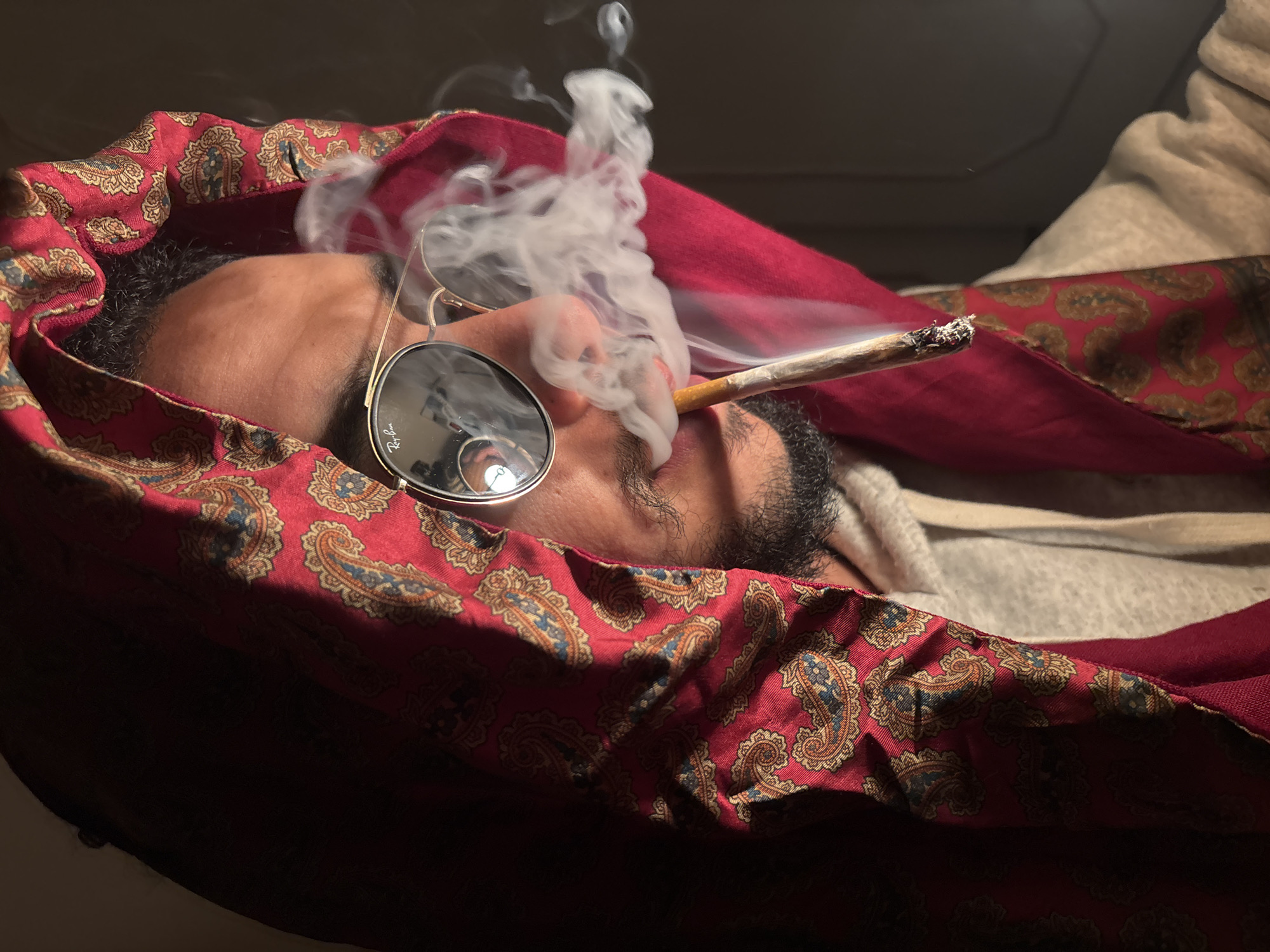 The image depicts a close-up, dimly lit, head-and-shoulders portrait of an individual from the left profile. The subject is an adult male with dark skin and a short beard and mustache. He wears mirrored aviator-style sunglasses with "Ray-Ban" branding visible on the left lens. A red fabric with a gold and blue paisley pattern is draped over his head and shoulders, resembling a hood or head covering. Beneath this fabric, he wears a light grey hooded sweatshirt. A lit hand-rolled cigarette with visible ash is held in his mouth, from which a dense plume of white smoke emanates, particularly around his mouth and nose. The background is uniformly dark and devoid of features. The lighting is strong from the left, highlighting the subject's face and the patterned fabric, while the right side of the frame and the background are cast in shadow. No specific urban or geographical identifiers are present to confirm a location in Tanger, Morocco.