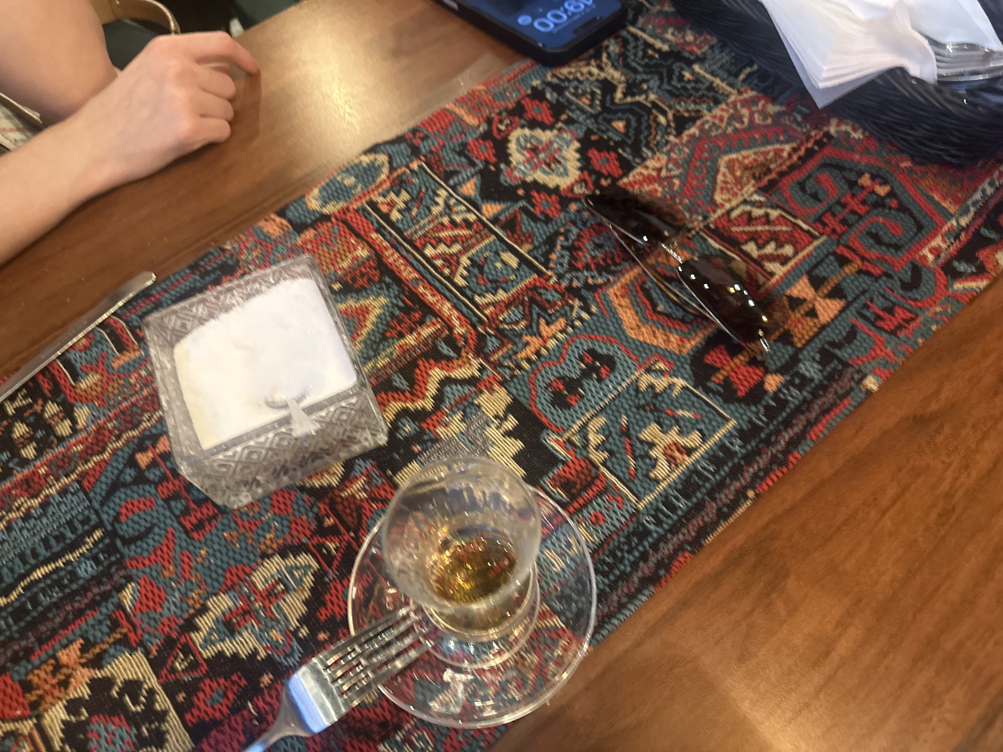 On a wooden table covered with a patterned runner of red, teal, black, and orange geometric shapes, a partial view of a person's arm rests on the left edge. A clear plastic container holding white napkins sits beside a silver knife. Further along the runner, sunglasses are placed on the pattern. A small glass cup filled with liquid is on a saucer with a fork lying beside it. Above the sunglasses, a black smartphone displays the time "19:00". To the right, a stack of white napkins are in a black plastic container. The scene is in the city of Unknown, Unknown.