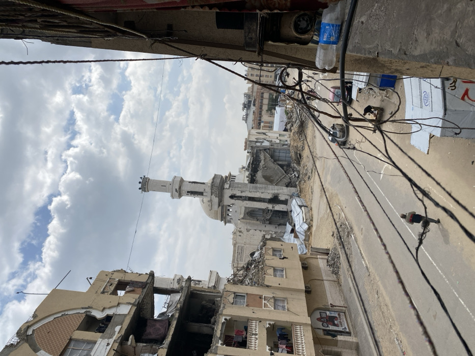The image captures a scene of widespread destruction in Khan Yunis, Palestinian Territory, under a cloudy sky. In the mid-ground, the severely damaged Great Mosque of Khan Yunis is prominent, with its minaret largely intact but the main building exhibiting significant structural collapse and exposed interior elements. Extensive rubble surrounds the mosque's base. To the left, a multi-story building, likely residential, shows severe damage with portions of its facade missing, revealing interior spaces and hanging laundry on balconies. A "SQUID GAME" graphic is visible on its lower exterior. The paved street below shows white lane markings, with two cars parked on the right side. Numerous black utility wires crisscross the foreground. A white plastic water bottle is hooked onto one of these wires on the far right. A male figure walks on the left side of the street, heading away from the viewer. Further down the street on the right, another male figure is bent over an object resembling a cart or stall, and additional smaller figures are visible in the distance. Piles of debris and twisted materials are present along the sides of the street, particularly near the damaged structures.