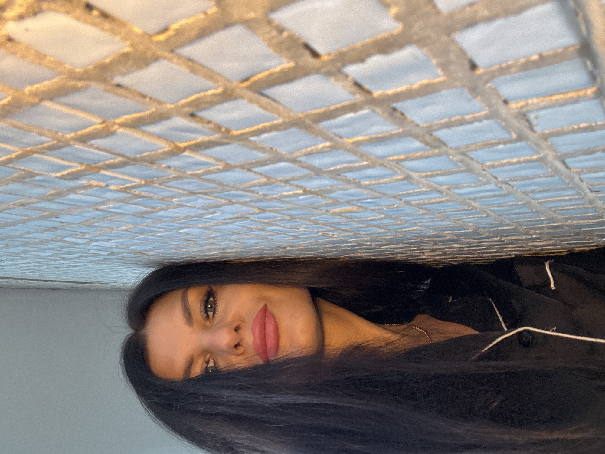 The image presents an upside-down view of a woman with long, dark hair, positioned beneath a tiled ceiling. The woman's face is visible, with her eyes open and lips appearing to be a light pink color. She wears a dark shirt or top with a white contrasting trim. Above her is a tiled ceiling consisting of small, square, light-colored tiles separated by a grid of darker grout. The location is indiscernible from the information provided.