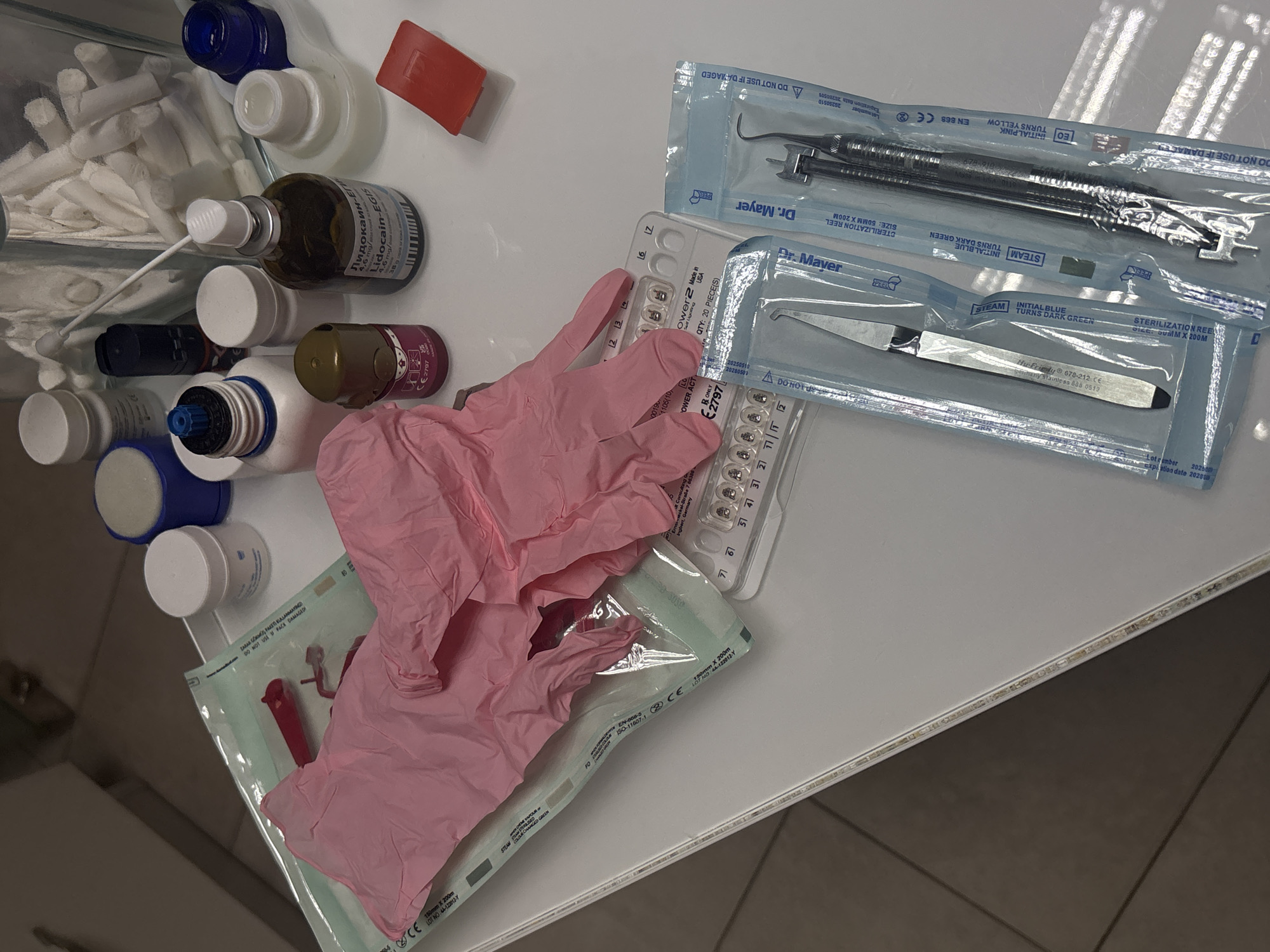 The image depicts a collection of medical supplies on a white surface, likely a countertop. Visible items include unopened sterile dental instruments packaged by "Dr. Mayer," pink disposable gloves still in their packaging, and a blister pack labeled "Power 2, Made in USA." There are also various bottles of medications, including one labeled "Lidocain-EGIS" in Russian. Cotton swabs and other small medical items are visible in a container. The focus is on sterile dental instruments and medications, suggesting a dental or medical environment. The city of Sofia, Bulgaria, is not determinable from the image.