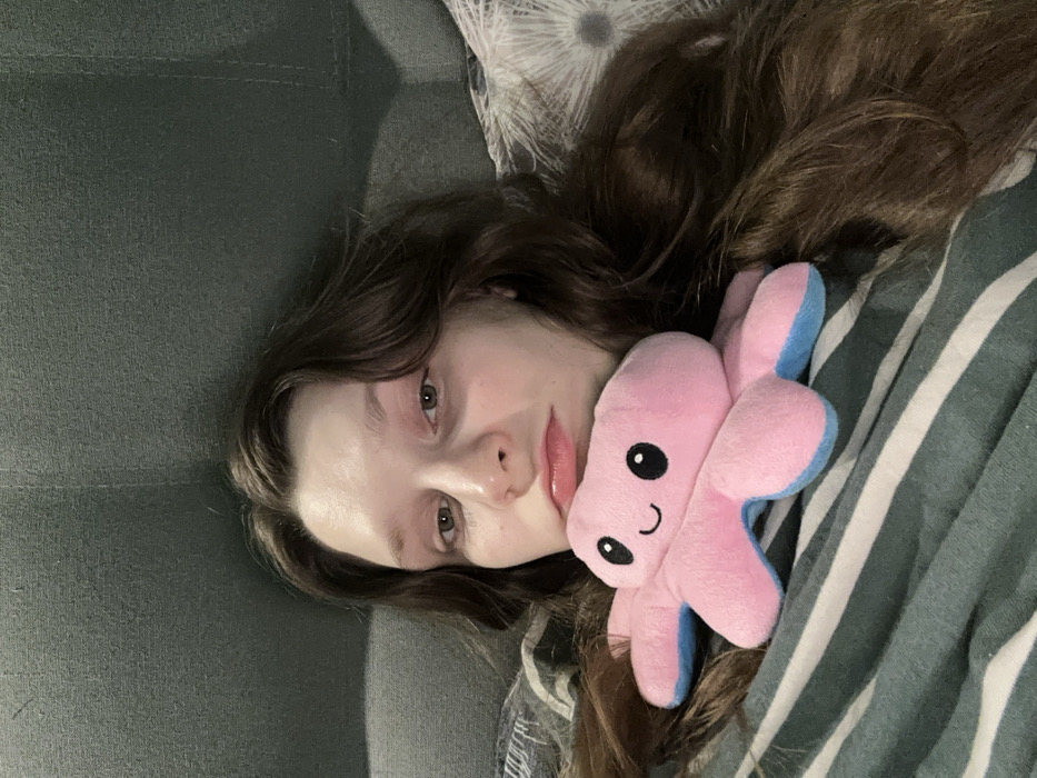 A young woman with long, wavy brown hair rests on a green striped blanket, a pink plush octopus with blue accents nestled close. She gazes towards the viewer with light eyes and glossy lips. Part of a green sofa is visible in the background, suggesting a relaxed setting. The scene is casually lit, creating a soft, intimate atmosphere.