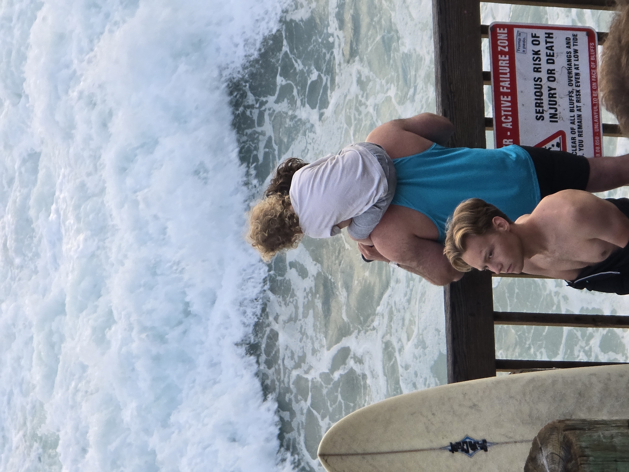 In Encinitas, California, USA, on a pier overlooking the Pacific Ocean, a man carrying a child on his shoulders faces the choppy water and waves. The man wears a turquoise tank top and black shorts. The child, with curly blonde hair, wears a white long-sleeved shirt and grey pants. To the left of the man, a light-colored surfboard stands upright against the wooden railing. In the foreground, a young man with blonde hair, shirtless and wearing black shorts, looks to the right. Behind him and to the right, a red and white sign is attached to the pier railing. The sign reads "R - ACTIVE FAILURE ZONE SERIOUS RISK OF INJURY OR DEATH. CLEAR OF ALL BLUFFS, OVERHANGS AND YOU REMAIN AT RISK EVEN AT LOW TIDE. 3.08.050 - UNLAWFUL TO BE ON FACE OF BLUFFS." A smaller sign above the larger one says "Trump is a pedo." The ocean is a turbulent blue-green with white foam.