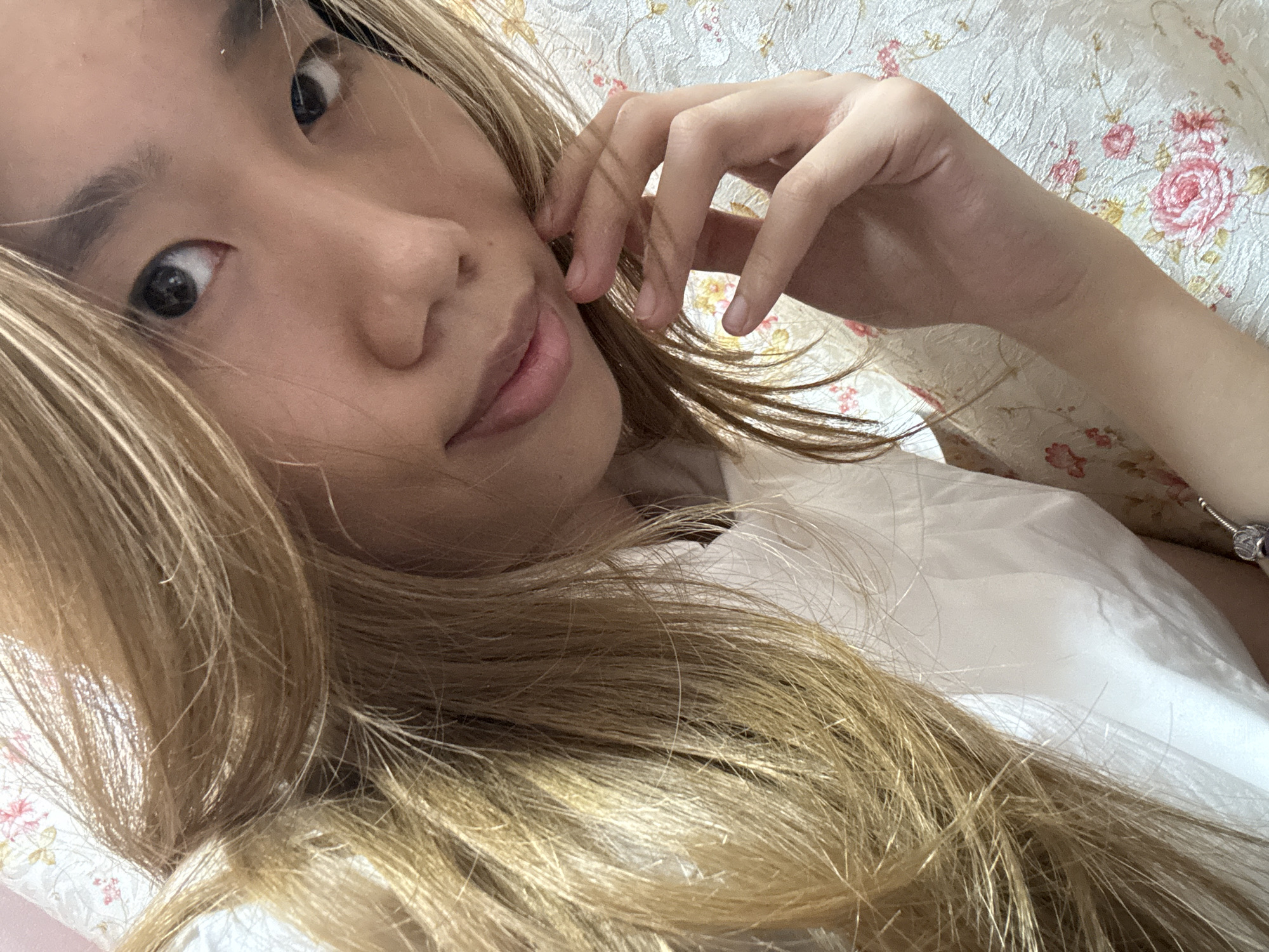 A young Asian woman with long, blonde, wavy hair is shown in a close-up selfie. Her hair is styled to fall over her right shoulder, framing her face. She is wearing a white collared shirt. Her left hand is raised, with her fingers gently touching her cheek. Her eyes are dark and looking directly at the camera. The background appears to be a floral-patterned fabric, possibly a bedspread or upholstery. The lighting is soft, suggesting an indoor setting. There are no discernible environmental features indicating a specific location within Shapingba District, China.